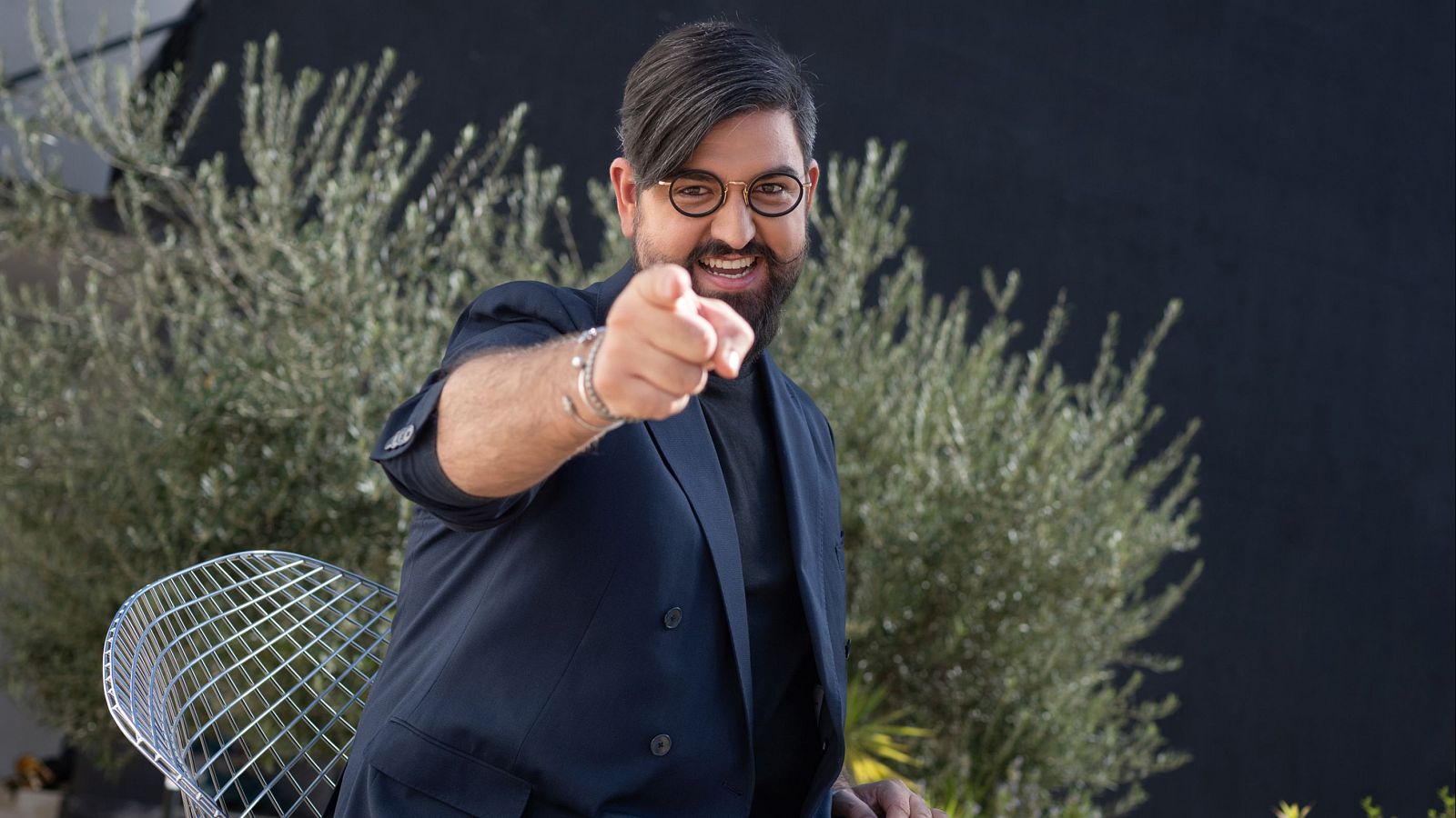 Un hombre con barba, gafas y chaqueta azul oscura sonríe y apunta a la cámara. Se encuentra frente a vegetación y una silla de metal.
