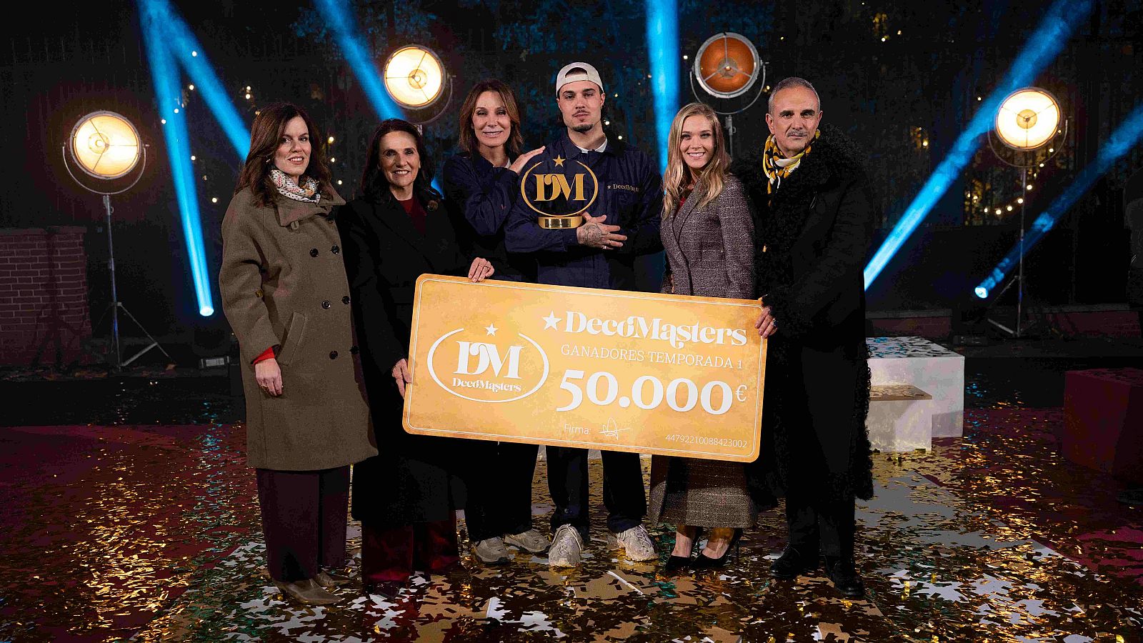 Mar Flores y Carlo Costanzia, ganadores, sostienen un cheque gigante en un escenario con focos y confeti. El hombre lleva un mono azul oscuro y gorra, mientras que la mujer está a su lado, rodeados de otras personas.