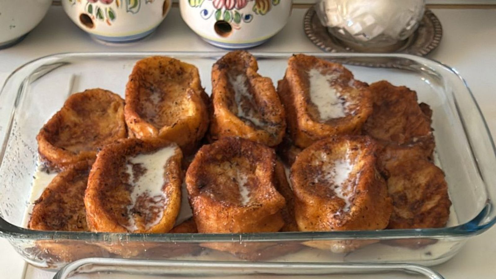 Un recipiente de vidrio rectangular contiene torrijas doradas, con cobertura cremosa y canela, sumergidas en leche. La disposición ordenada sugiere una preparación cuidadosa.