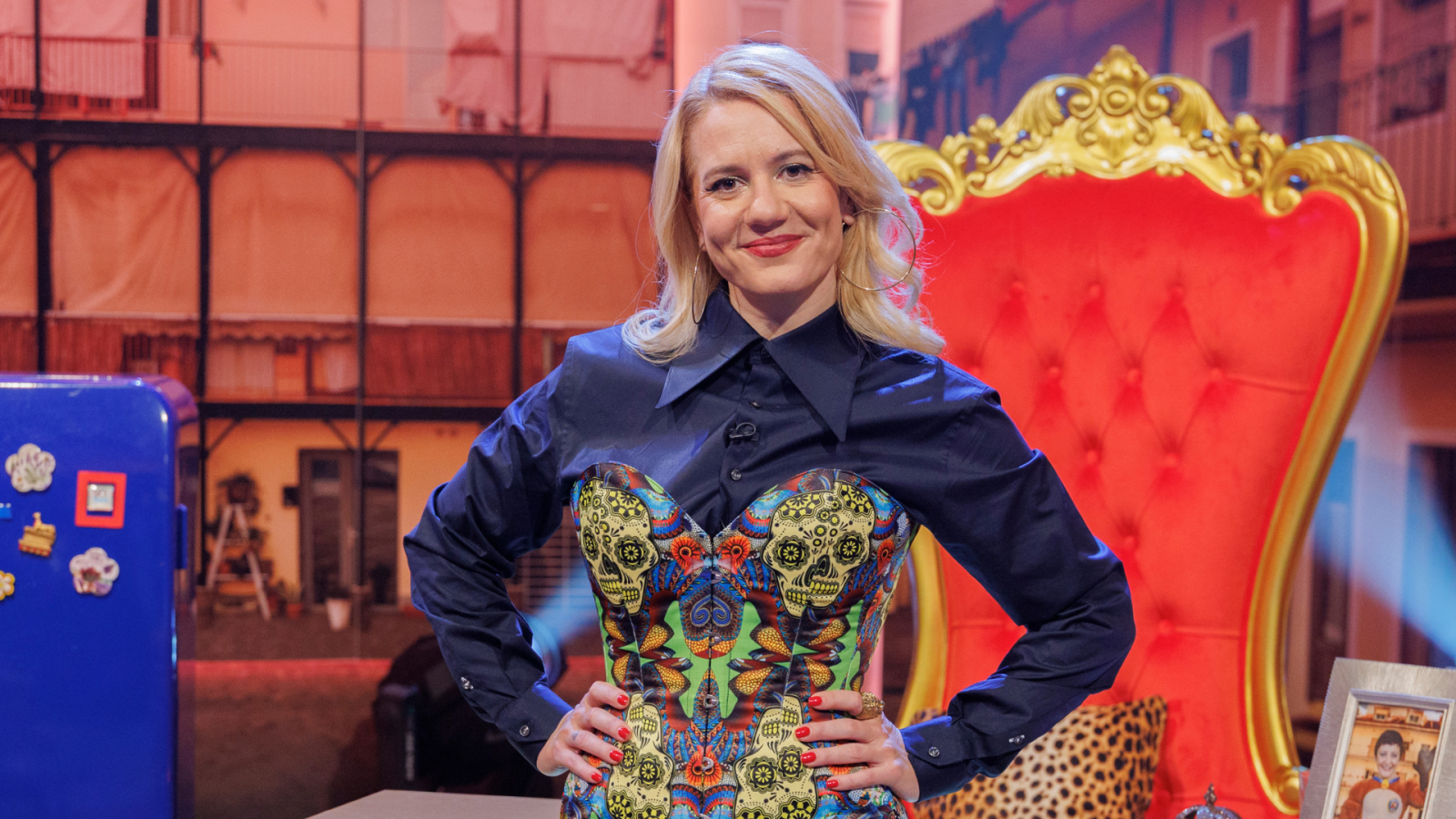 Henar Álvarez, presentadora de televisión, luce una camisa azul oscuro sobre un corsé estampado con calaveras, posando frente a una silla roja con detalles dorados, en un decorado urbano.