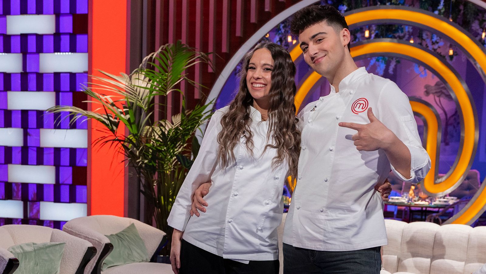 En un estudio televisivo, Masi Rodríguez y Juanjo Bona, vestidos con chaquetas de chef blancas con el logo de 'MasterChef', sonríen y posan para la cámara. Juanjo Bona señala el logo.