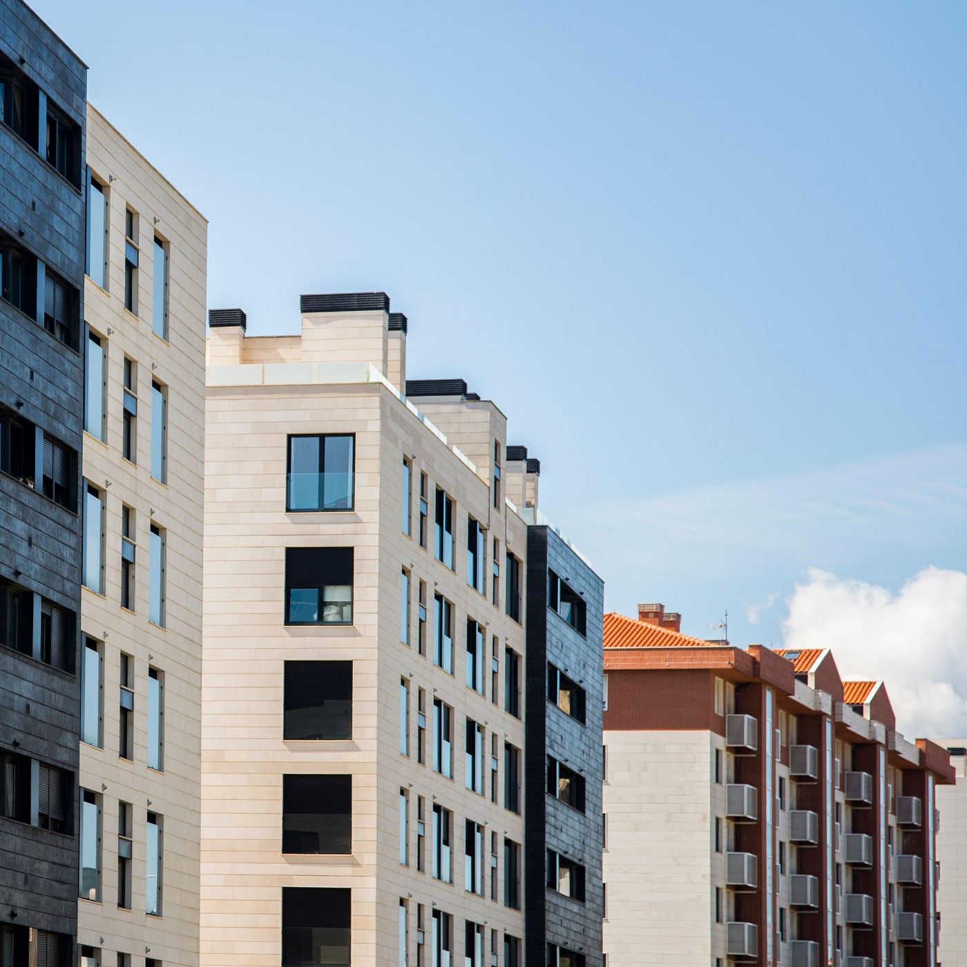 Una vista aérea muestra diversos edificios de apartamentos con fachadas de diferentes colores y diseños, incluyendo tonos grises, crema y beige, con ventanas rectangulares y algunos detalles en rojo y marrón, contra un cielo azul claro.