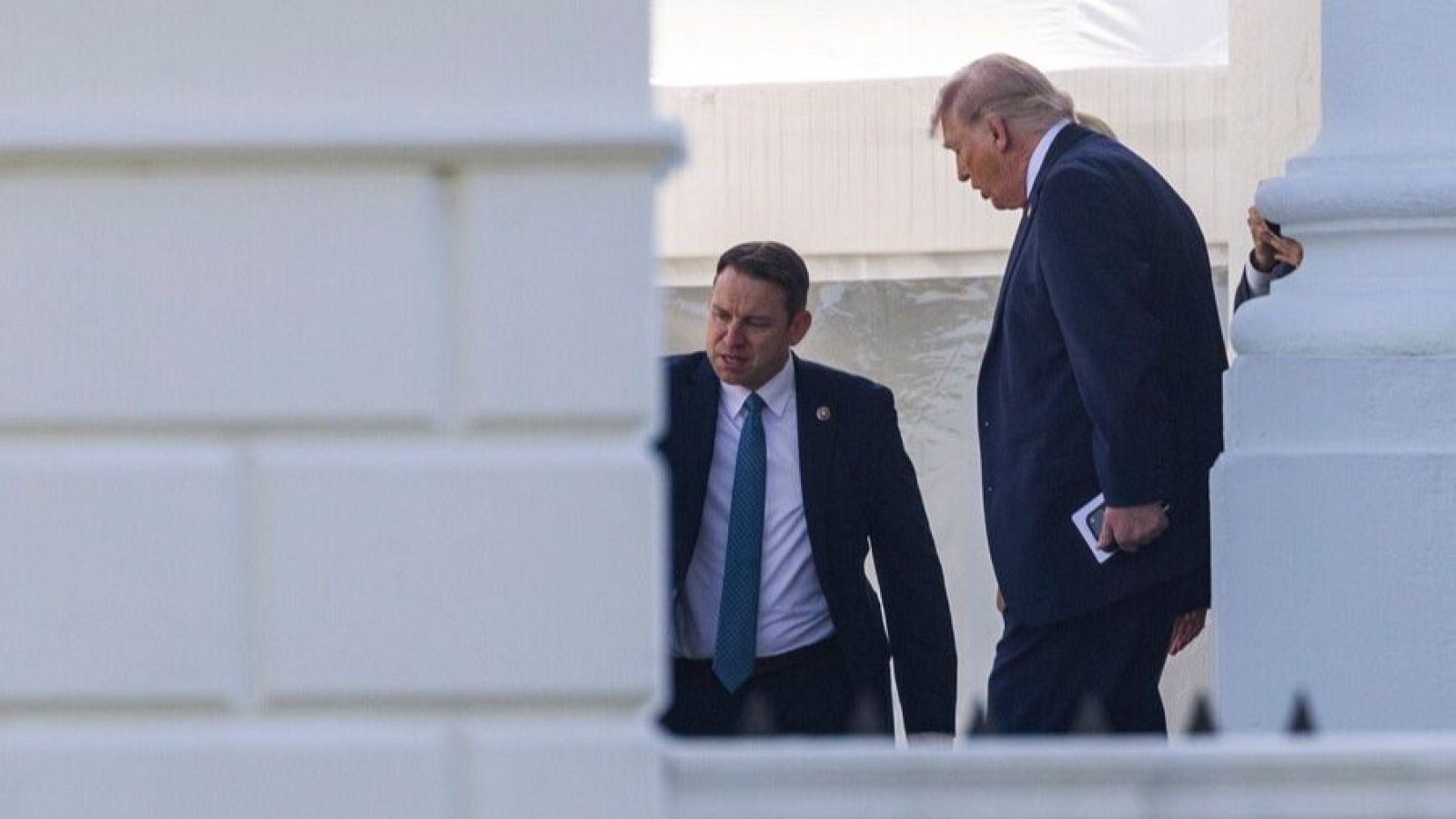 Donald Trump, vestido con traje oscuro, se dirige hacia un coche, acompañado por un hombre también vestido de traje. La escena transcurre en el exterior de un edificio con columnas.