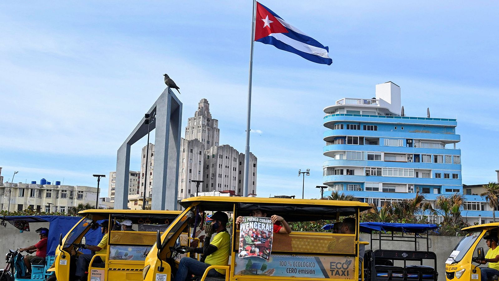 Una calle cubana muestra un monumento con una paloma y taxis eléctricos amarillos. Un conductor exhibe un cartel con la imagen de un líder político y la frase "¡El pueblo cubano vencerá!", en un contexto de crisis energética.