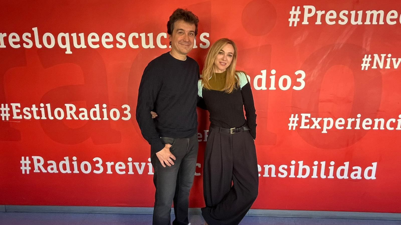 Un hombre y una mujer se encuentran de pie frente a un fondo rojo con hashtags. El hombre lleva un jersey oscuro y vaqueros, mientras que la mujer viste una blusa negra y pantalones anchos oscuros.