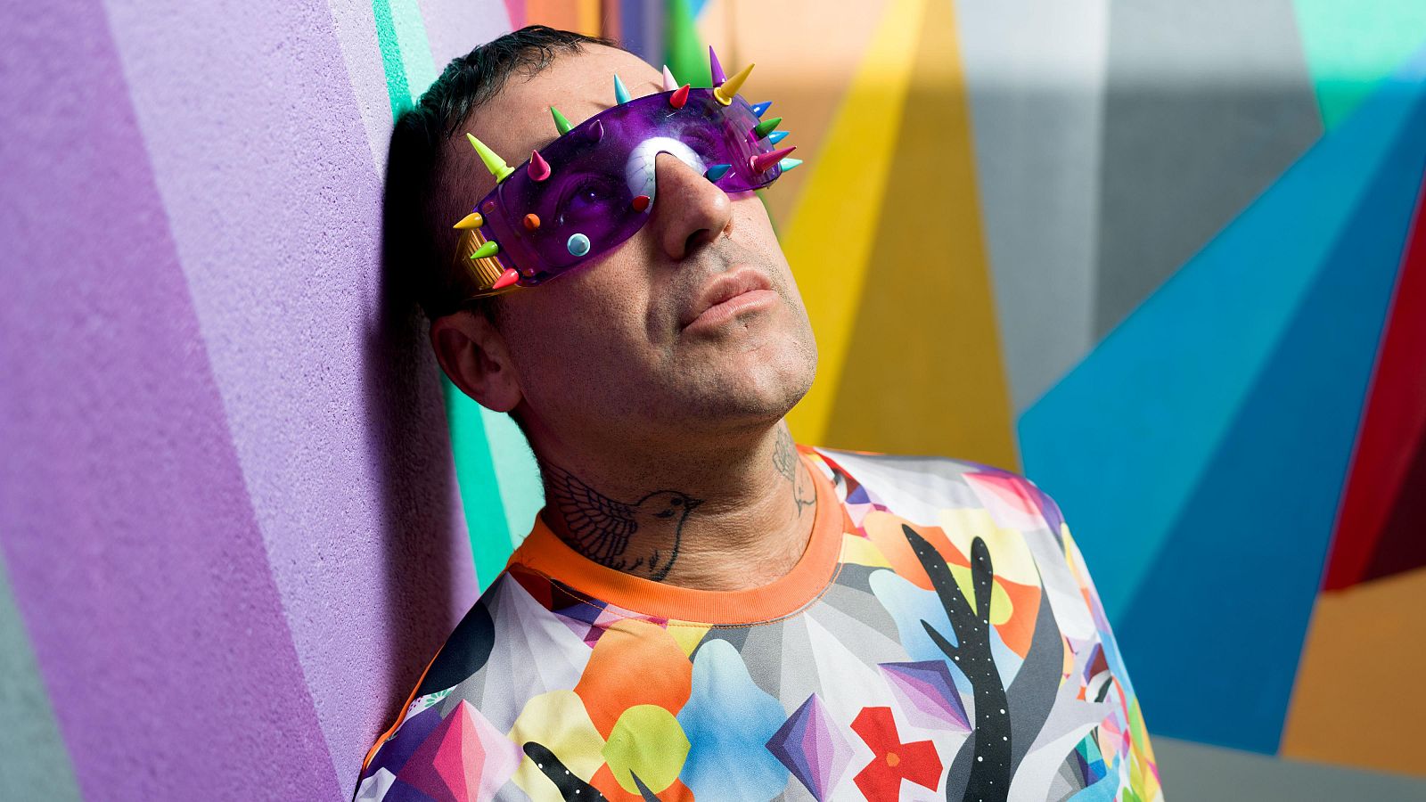 Okuda San Miguel, con gafas de sol y un tatuaje de pájaro, posa frente a una pared rayada. Viste una camiseta estampada con diseños geométricos y el fondo presenta formas abstractas en colores brillantes.
