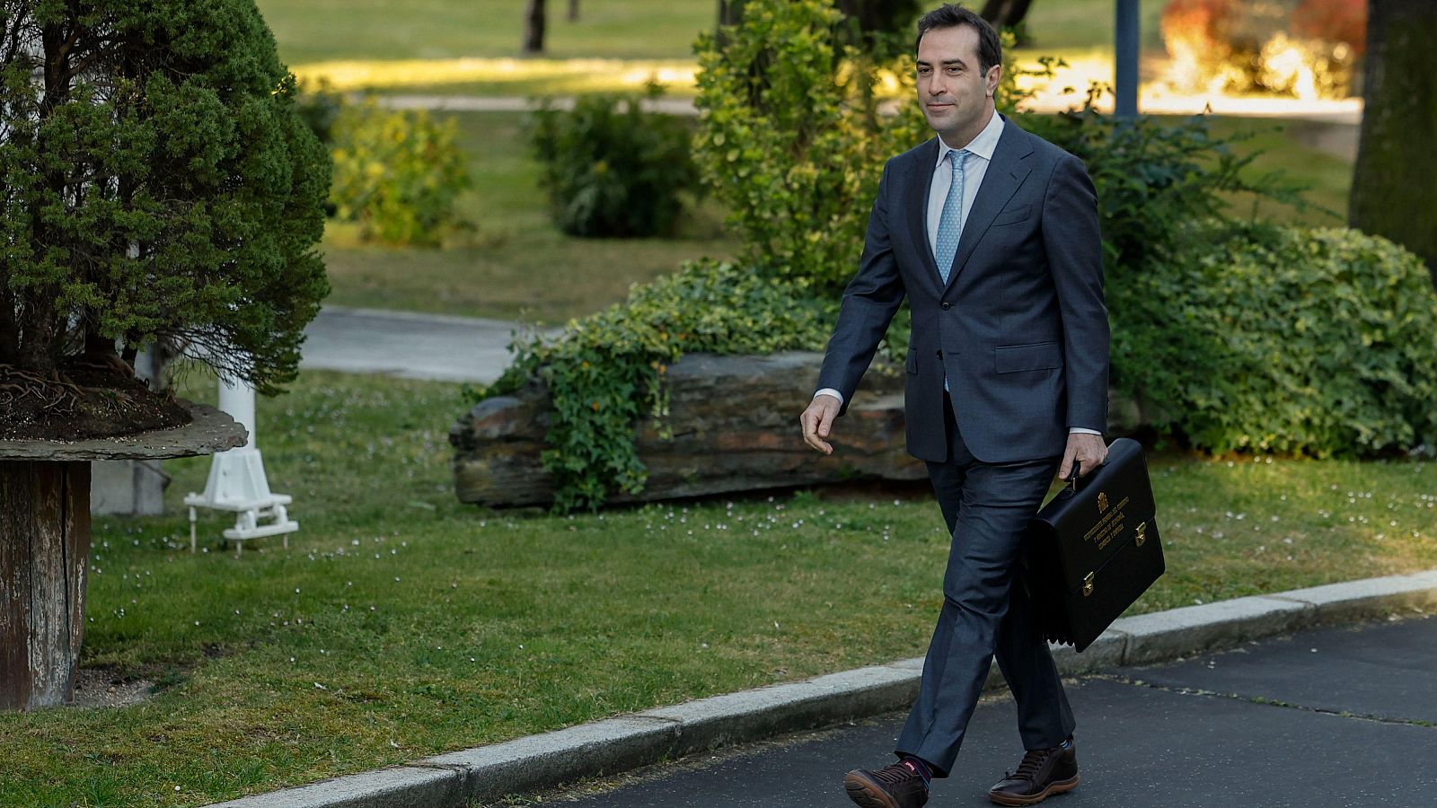 Carlos Cuerpo, ministro de Economía, camina al aire libre con un traje gris oscuro, camisa blanca y corbata celeste, portando un maletín negro. El fondo muestra vegetación y un camino.