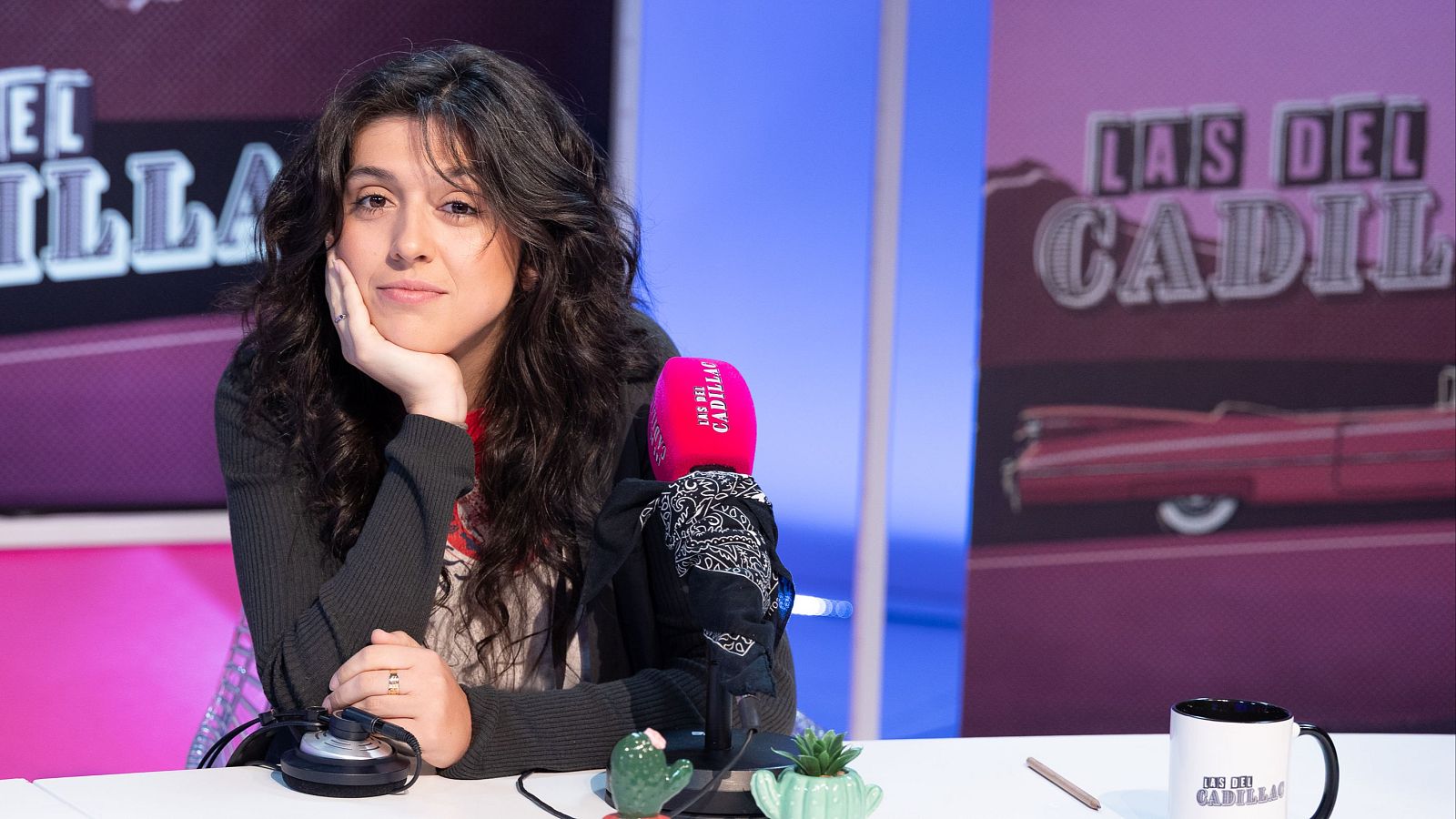 Claudia Polo en "Las del Cadillac", con micrófono rosa, auriculares y taza del programa. Fondo morado con logo y coche clásico.