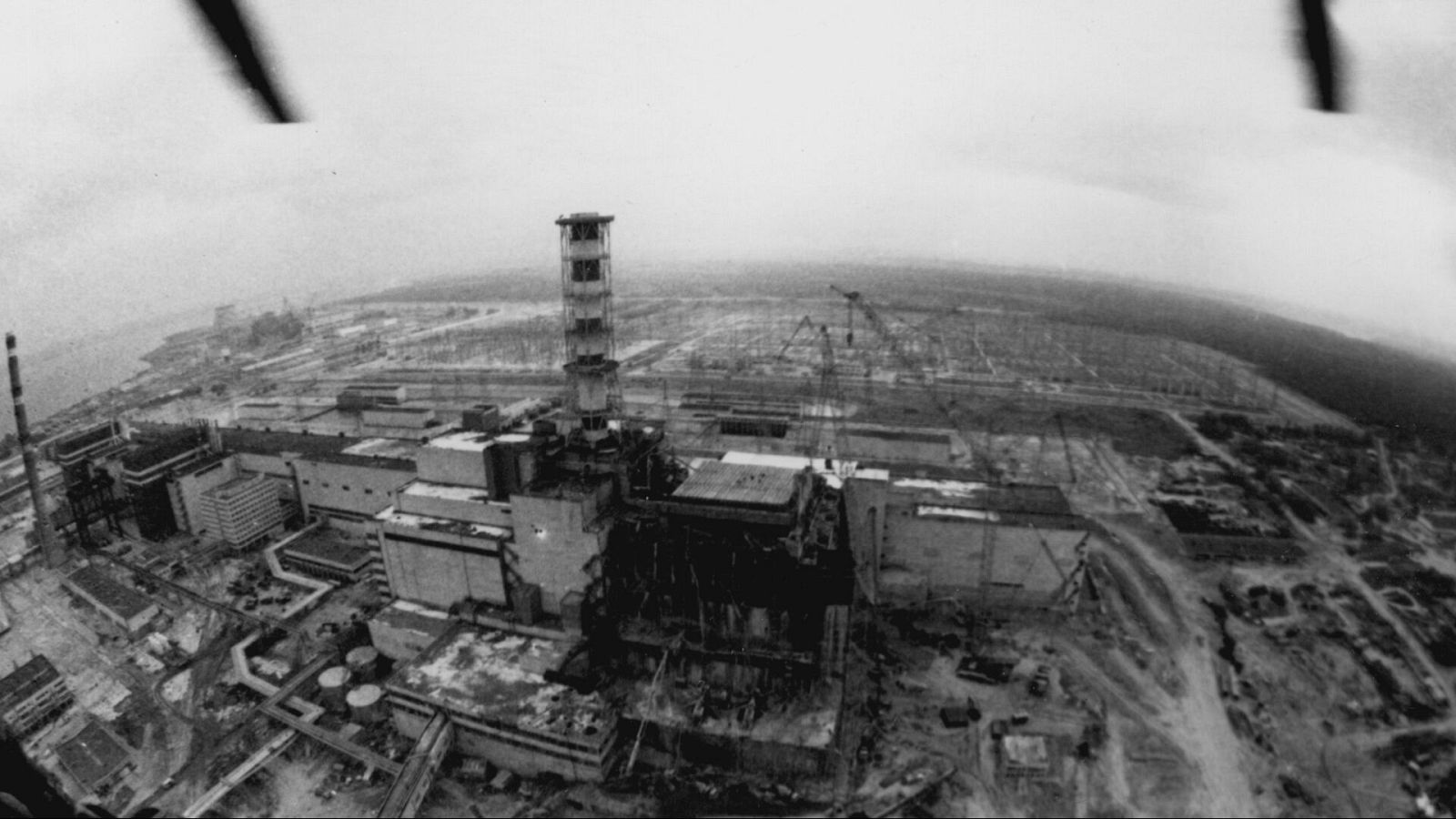 Vista aérea en blanco y negro de una central nuclear dañada, con escombros y estructuras afectadas. La imagen transmite una sensación de devastación y desolación, mostrando una zona desolada con vegetación escasa.