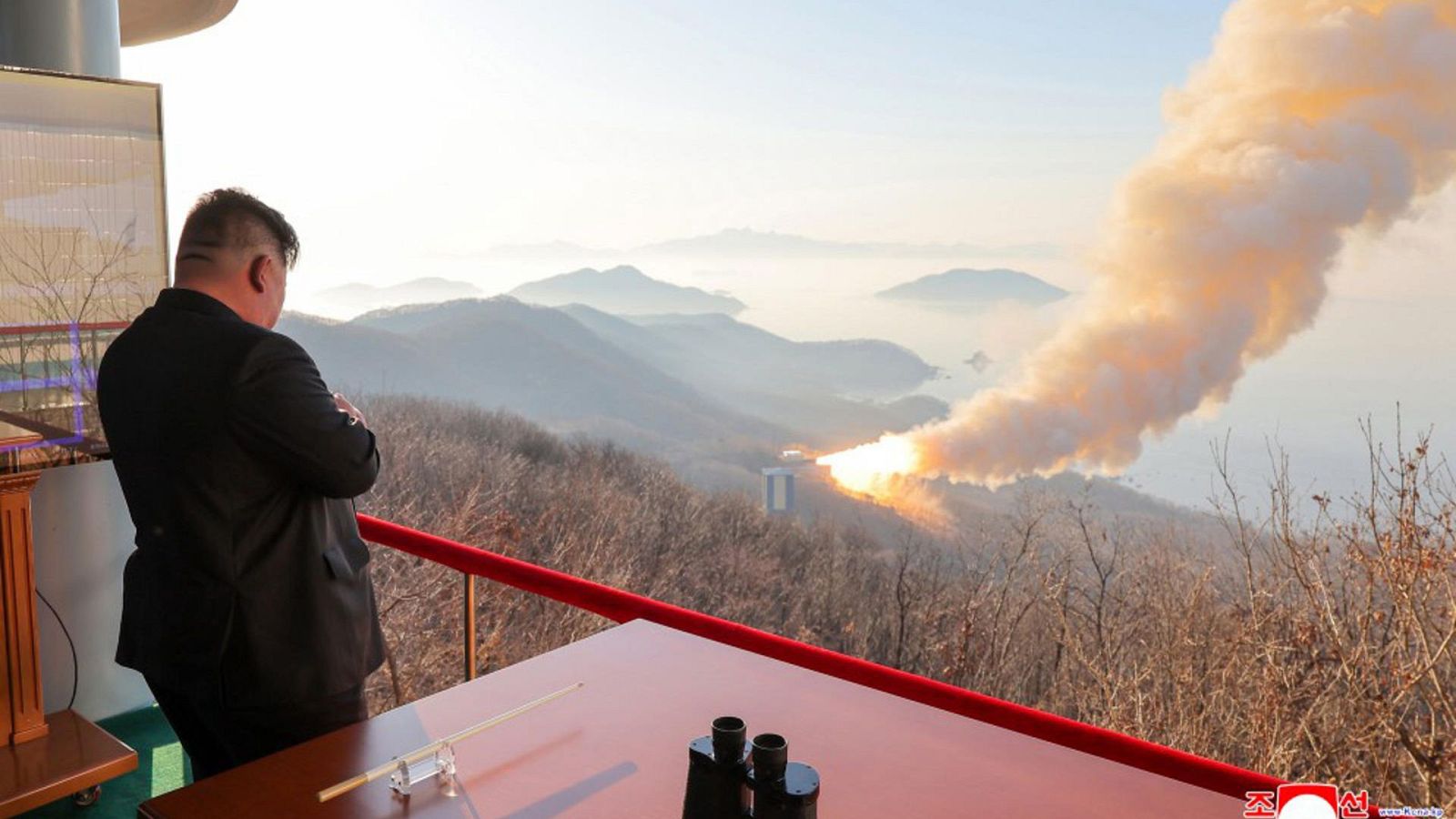 Fotografía que muestra al lider norcoreano, Kim Jong-un, supervisando una prueba de propulsión de misiles desde un balcón desde el que se ve un lanzamisiles echando humo en la ladera de un monte