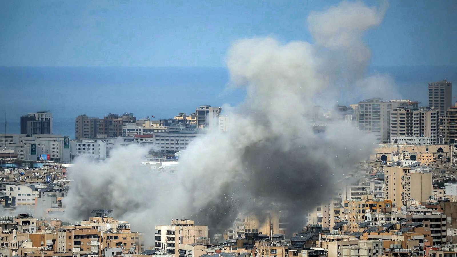 Imagen aérea de una zona urbana con edificios de diversas alturas y estilos. Una columna de humo denso se alza desde el centro, posiblemente indicando una explosión. El mar se aprecia al fondo.
