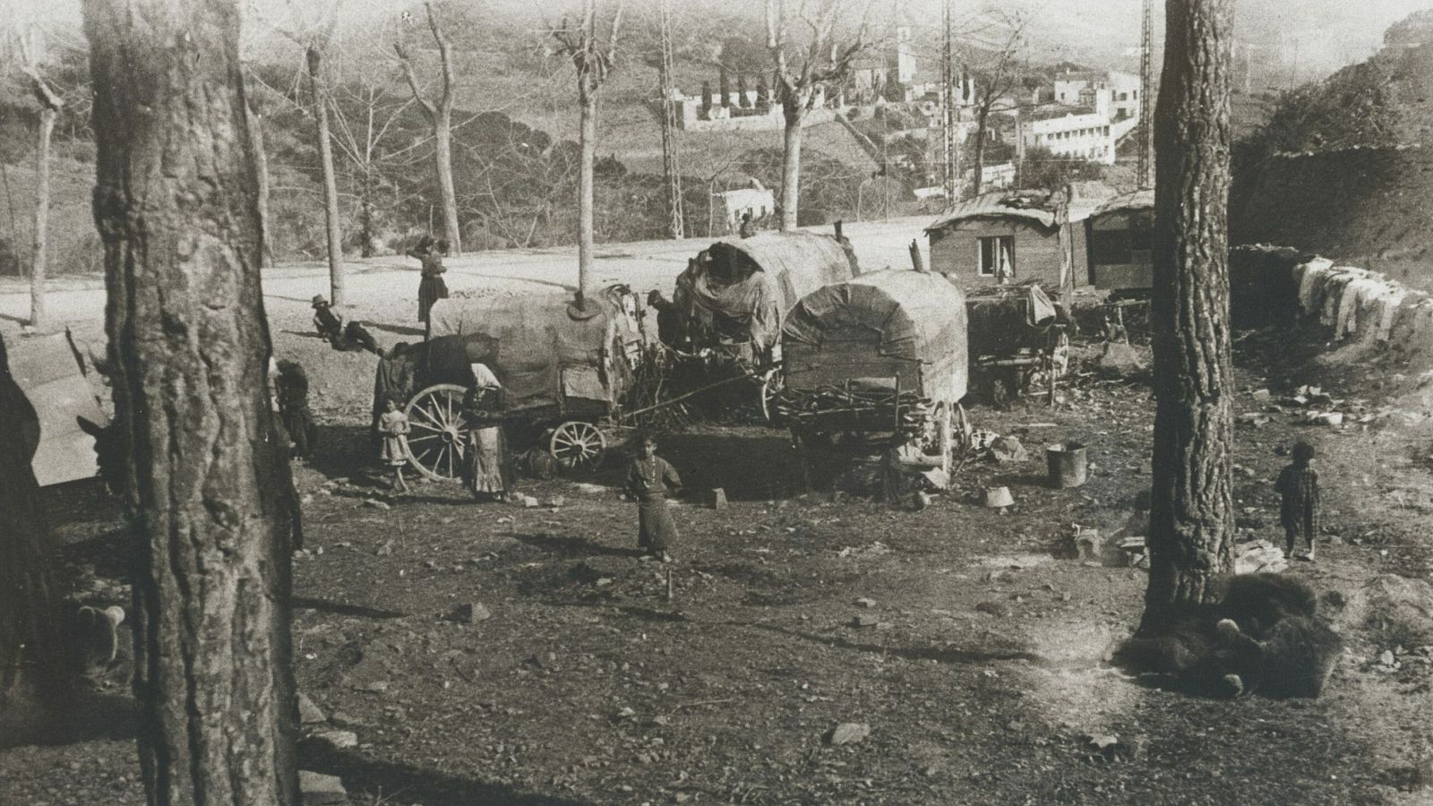 Una imagen en blanco y negro muestra una caravana con carromatos cubiertos y personas realizando actividades en un entorno rural, sugiriendo una vida nómada.