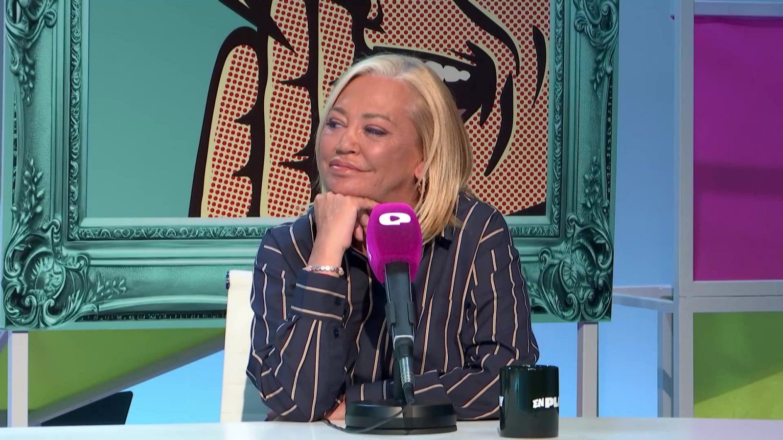 En la imagen, una mujer rubia con una camisa a rayas está sentada en una mesa, apoyando la barbilla en su mano. Frente a ella, se observa un micrófono morado y una taza negra.