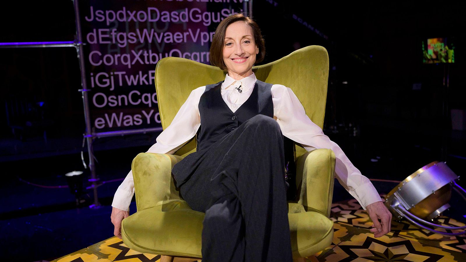 Una dona, vestida amb camisa i armilla, està asseguda en un sofà verd, amb les cames creuades i somrient. Al fons, es distingeix una estructura metàl·lica i un objecte amb llums.