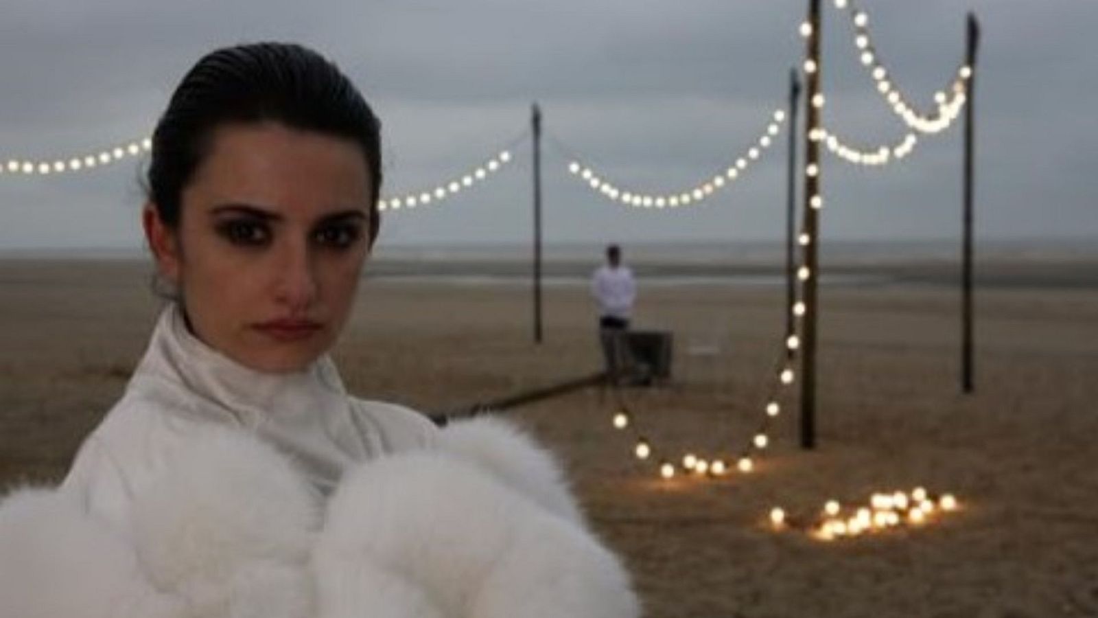 En primer plano, Penélope Cruz luce un abrigo blanco de piel con una expresión seria, en una playa con iluminación tenue, donde se observa una figura a lo lejos y una estructura con luces.