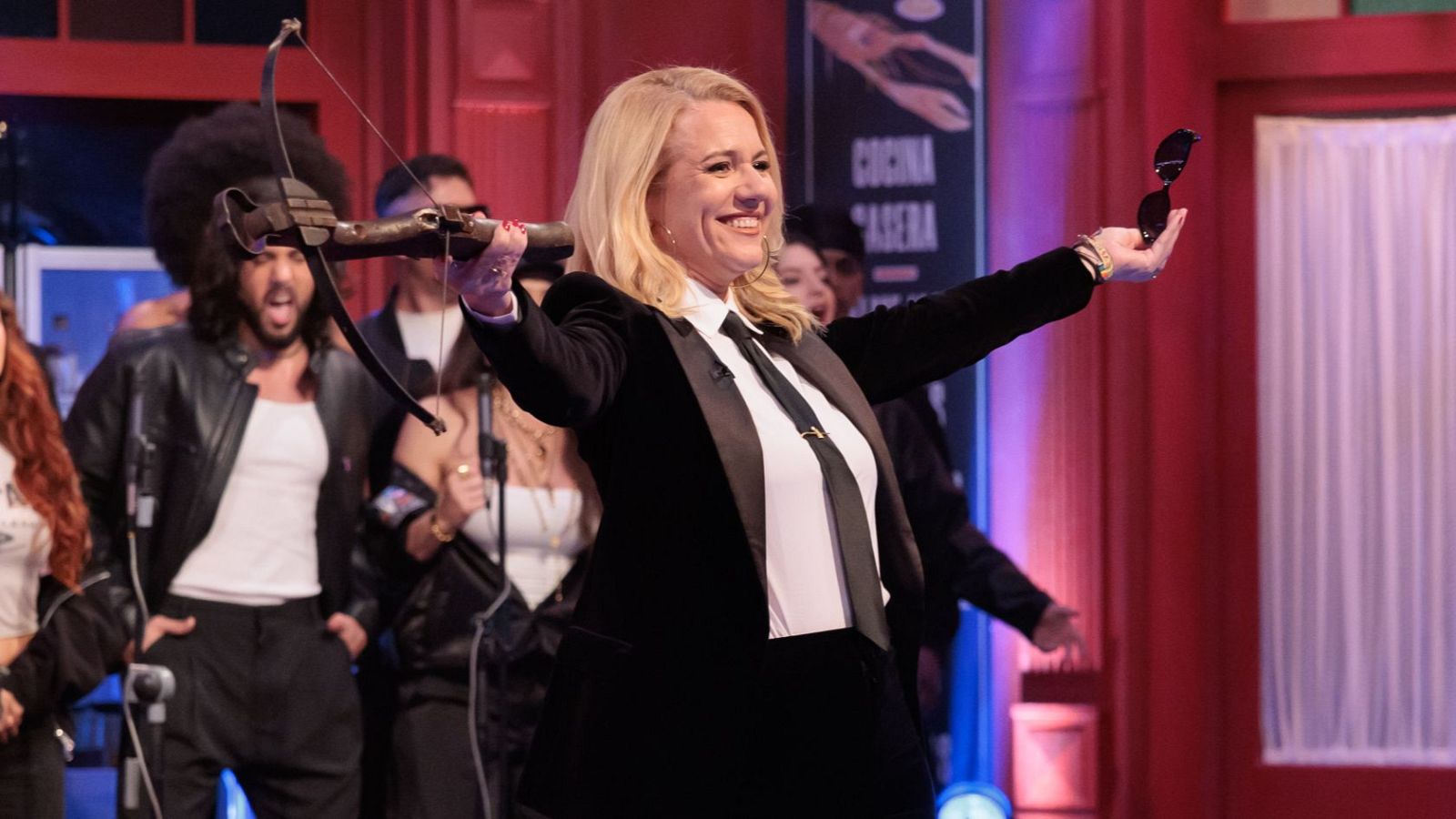 Henar Álvarez, sonriente, posa con una ballesta y gafas de sol, vestida con traje negro y camisa blanca. Detrás, un escenario con luces y decorados, y un hombre con afro.