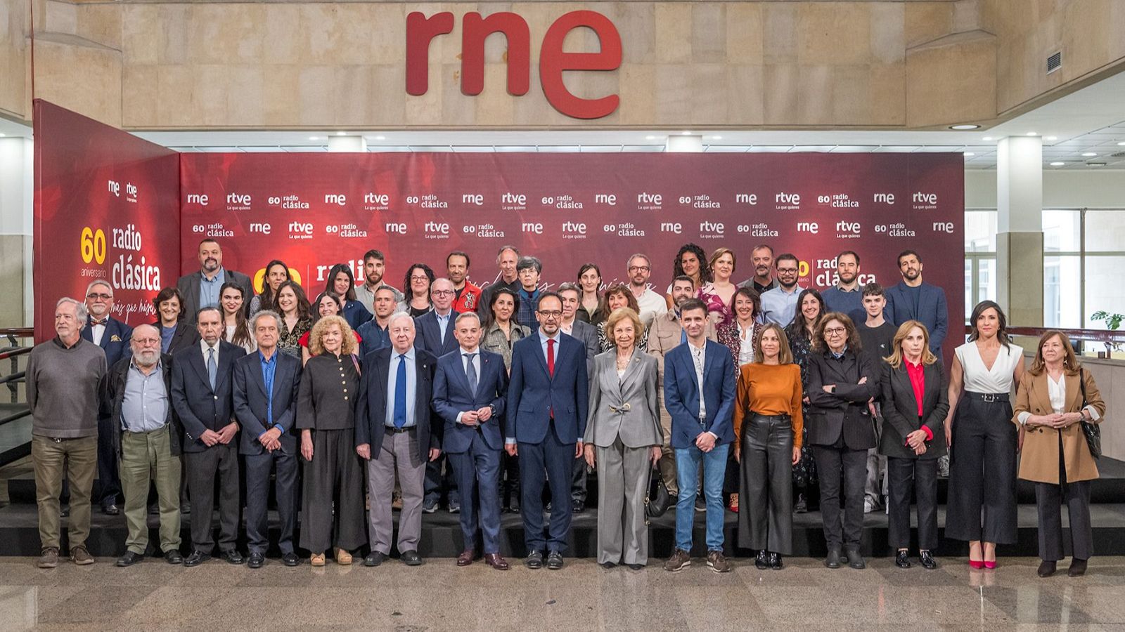 La Reina Sofía, vestida con un traje de chaqueta, posa para una fotografía de grupo en un evento conmemorativo. Se encuentra rodeada de personas, posiblemente trabajadores, frente a un panel con logotipos y la inscripción "60 Radio Clásica".