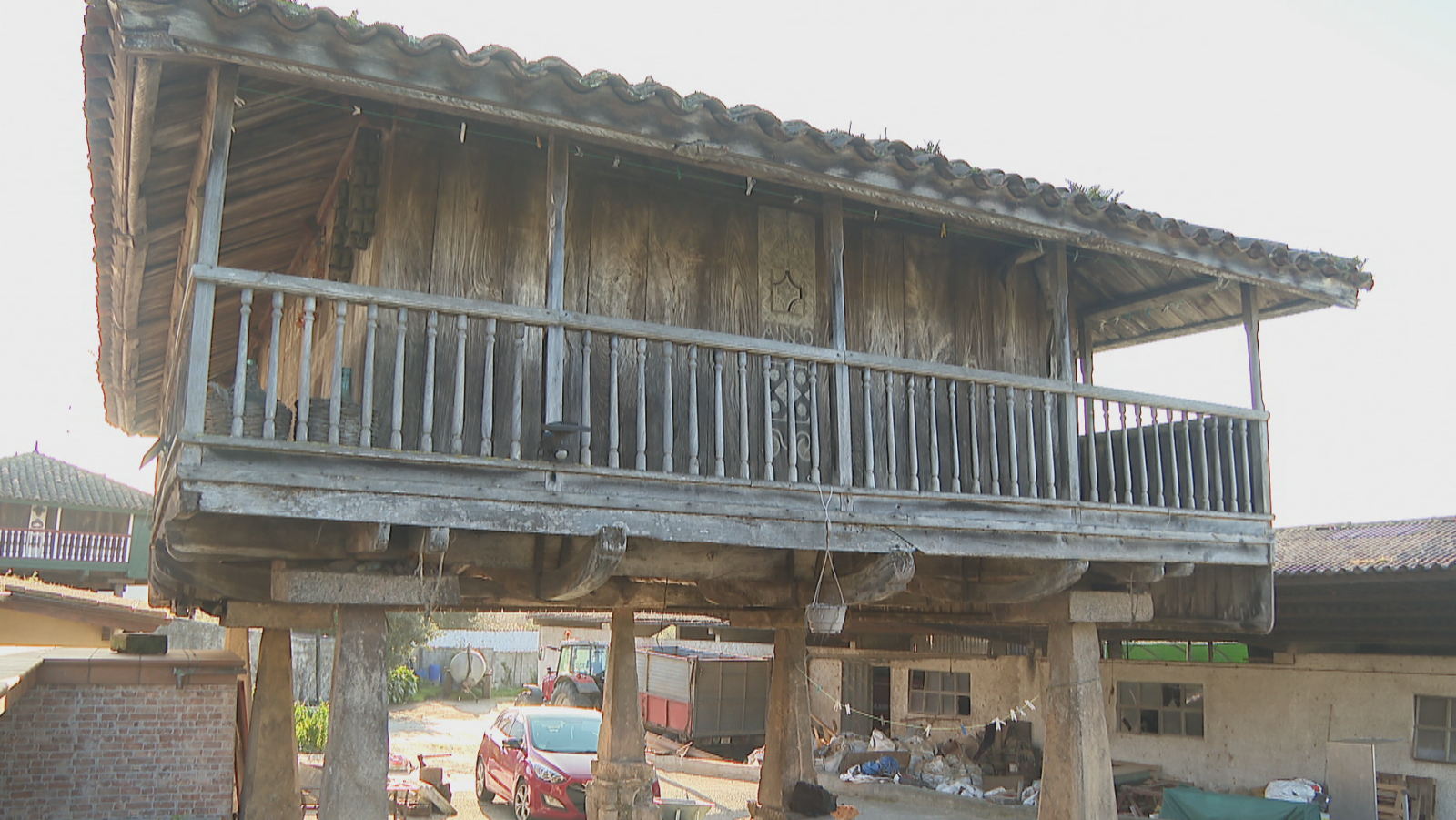Un hórreo de madera con tejado de tejas se alza sobre pilares de piedra. Presenta un balcón y detalles tallados en la fachada. En el entorno, se observan otros edificios, un coche, un tractor y una furgoneta.