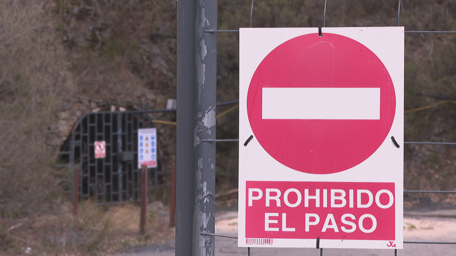 En la imagen se observa la entrada a una mina, con una señal de prohibido el paso en primer plano. La entrada está protegida por una puerta metálica con barrotes, y se aprecian otros carteles informativos.