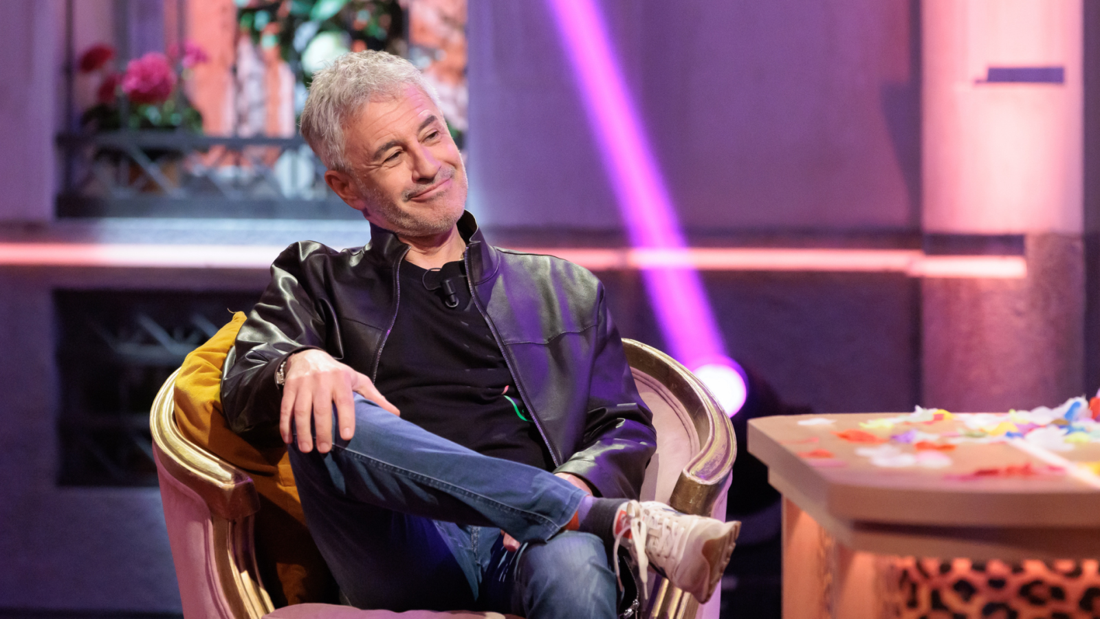 Sergio Dalma, con vestimenta casual, posa sonriente en un sillón. El fondo muestra una decoración floral y una mesa baja con confeti.