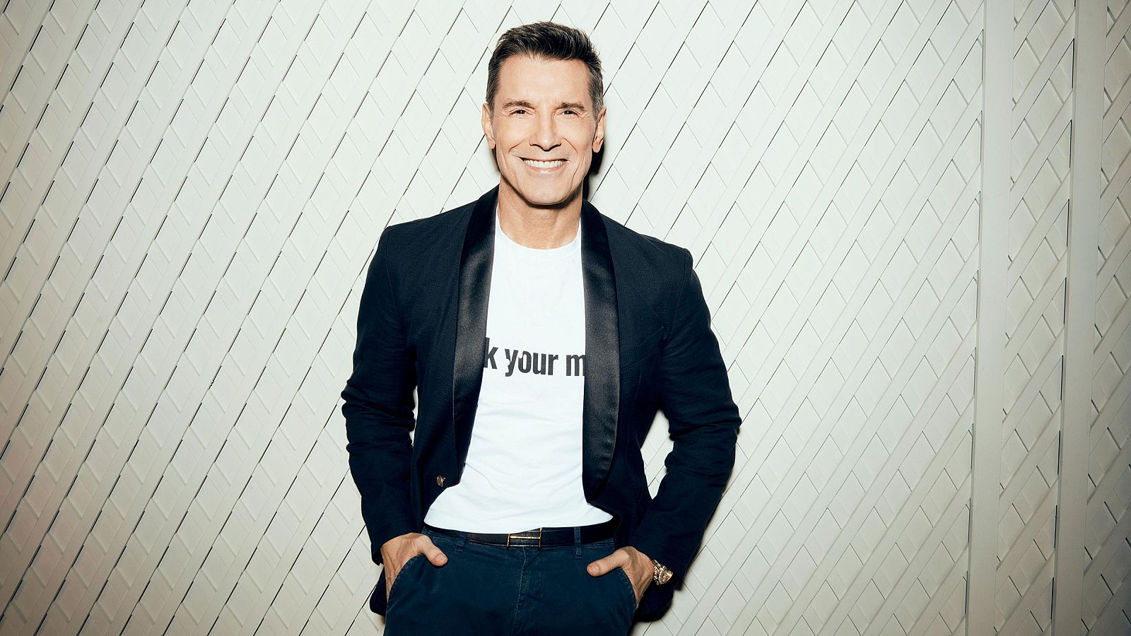 Jesús Vázquez, con una sonrisa, viste una camiseta con letras y una chaqueta negra. Sus pantalones oscuros y un cinturón con hebilla dorada completan su look, con un reloj dorado visible. El fondo es una pared blanca con relieve.