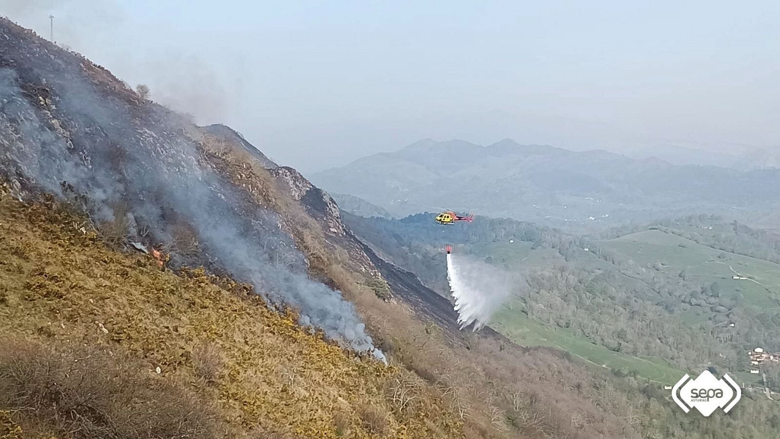 Un helicóptero vuela sobre un incendio forestal en una zona montañosa, lanzando agua para combatir las llamas y el humo que asciende.
