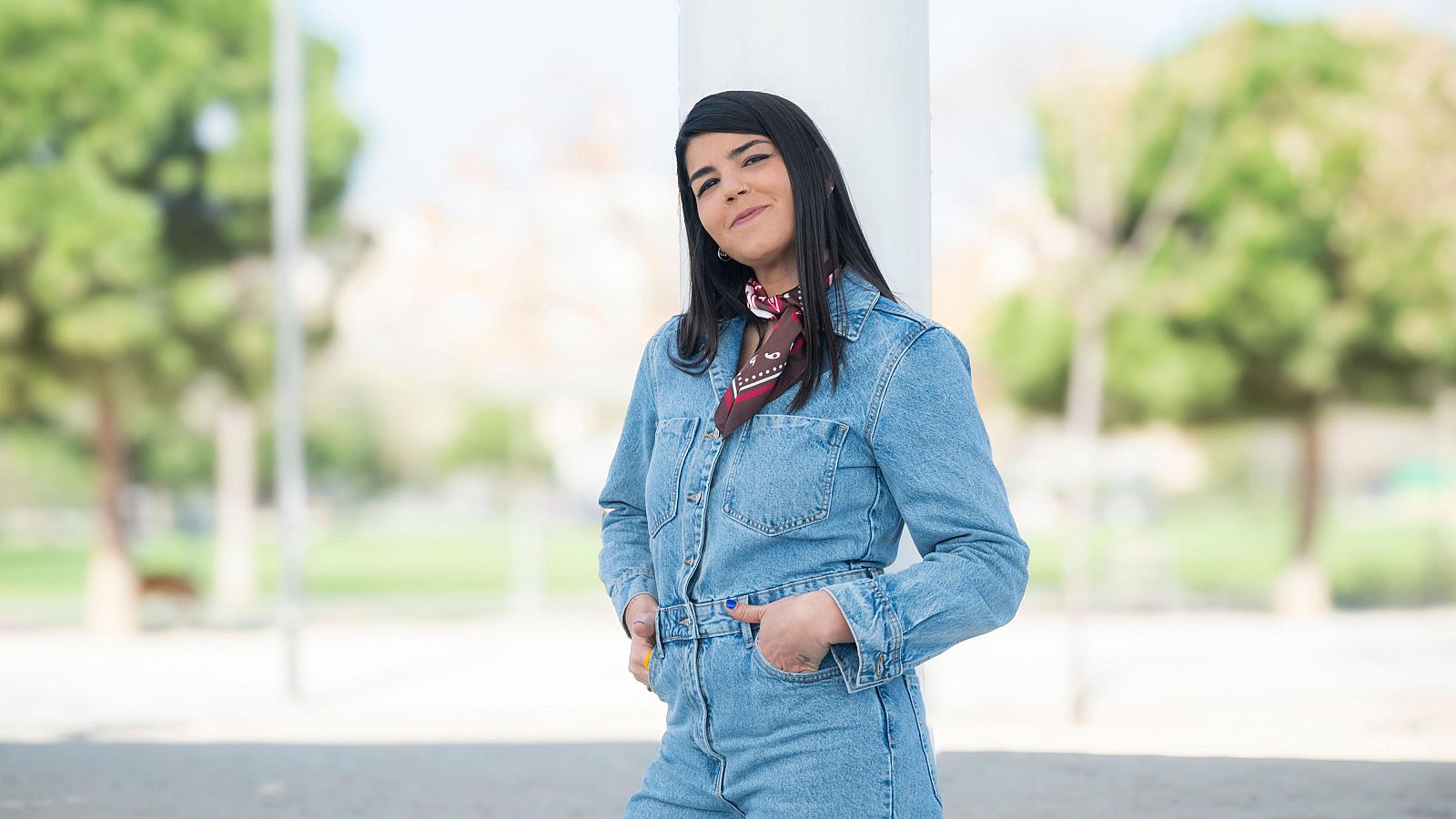 Una dona amb cabells negres i pell clara, vestida amb un conjunt vaquer blau clar i un mocador estampat, està recolzada en una columna blanca en un entorn exterior.