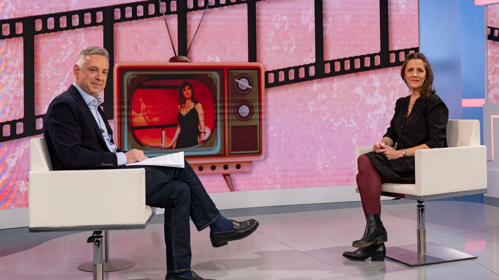 En un estudio televisivo, Jenaro Castro entrevista a una mujer vestida de negro. El presentador, con camisa clara y chaqueta oscura, sostiene papeles. El fondo muestra elementos que simulan un estudio de televisión.