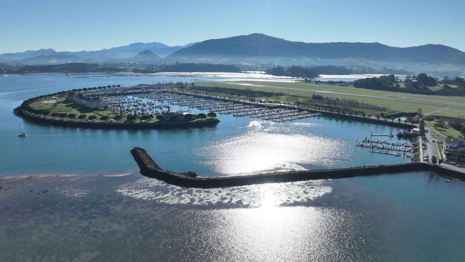 Una vista general de un puerto deportivo en una bahía de aguas tranquilas, con barcos atracados y montañas al fondo. El cielo es azul y despejado, y una escollera protege el puerto.