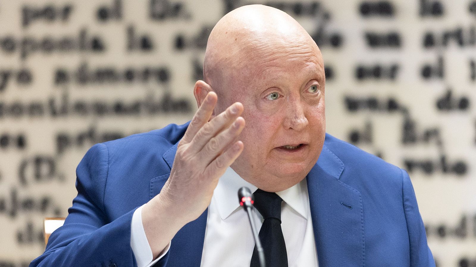 Fernando Samper, con chaqueta azul y corbata negra, gesticula frente a un micrófono. Se observa un texto borroso en el fondo.