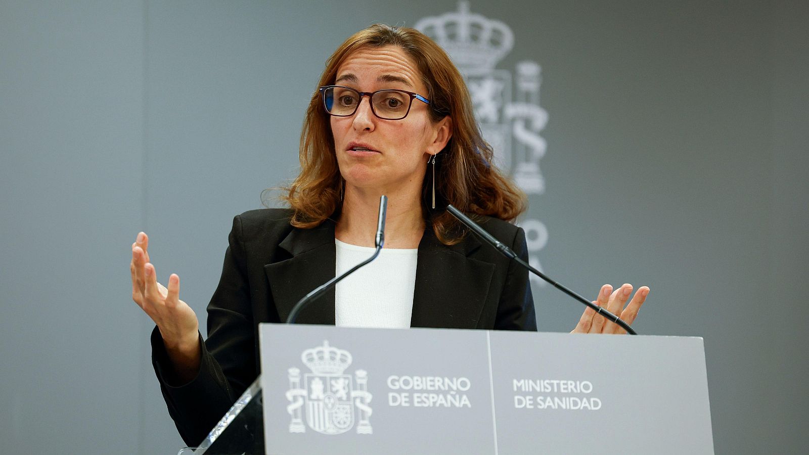 Una mujer con gafas y chaqueta negra, posiblemente Mónica García, habla desde un atril con los logos del Gobierno de España y el Ministerio de Sanidad.