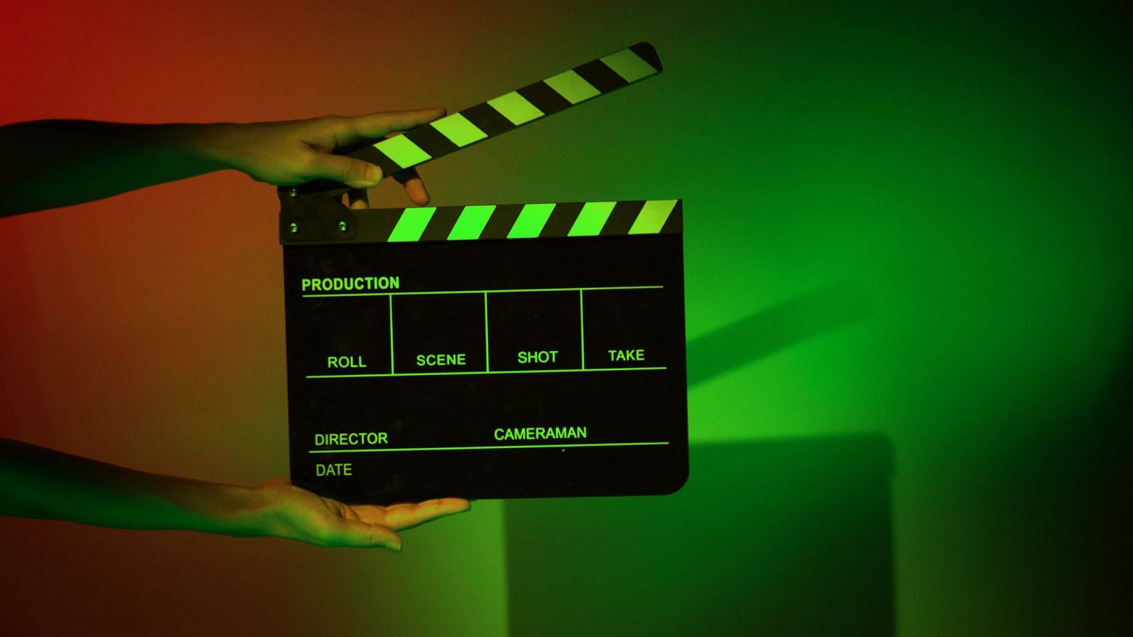 Una claqueta de cine negra con rayas verdes, abierta y sostenida por dos manos, muestra información relevante para la producción cinematográfica, con un fondo iluminado en rojo y verde.