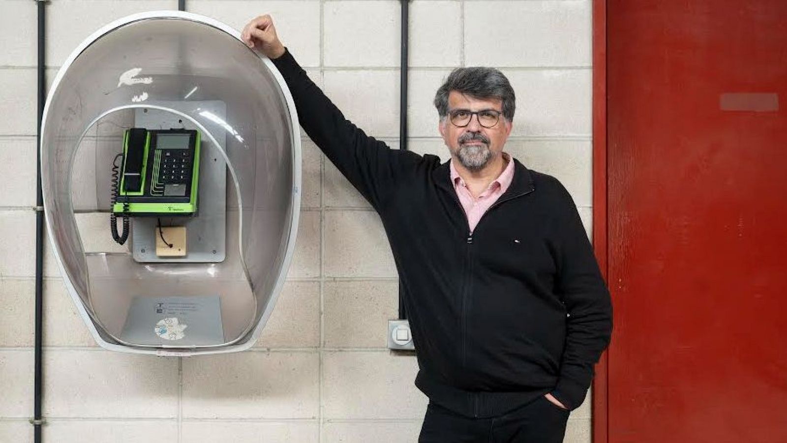 Un home amb barba i ulleres, vestit amb jaqueta negra i camisa rosa, està al costat d'una cabina telefònica moderna amb un telèfon verd. A la dreta, es veu una porta vermella.