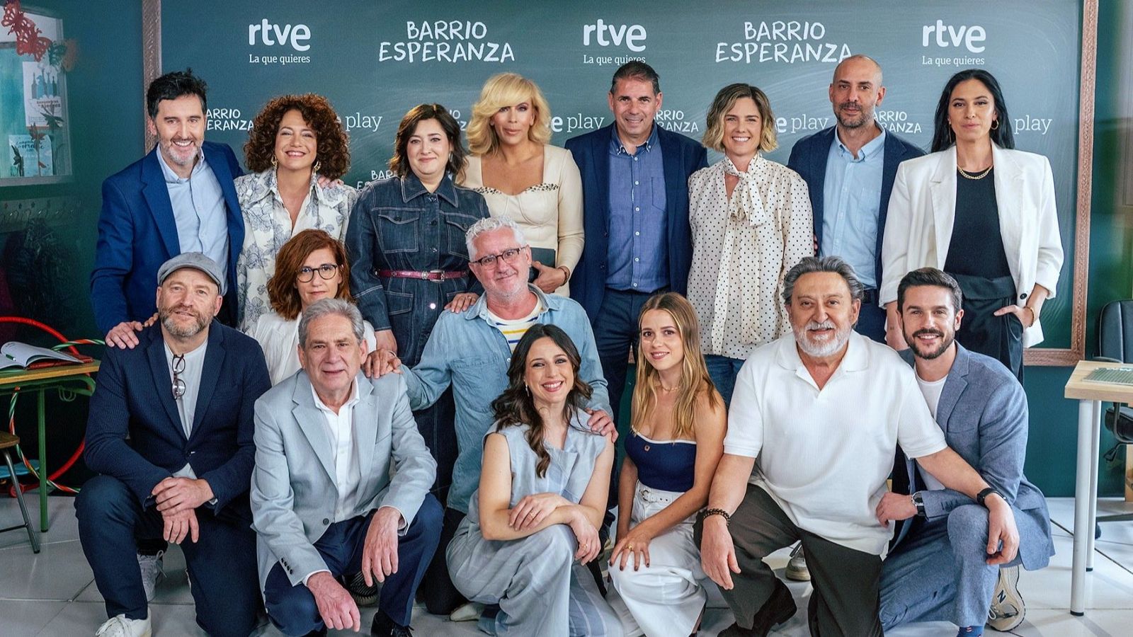 Un grupo de actores y actrices posa para una fotografía promocional frente a una pizarra con el logo de RTVE y el título de la serie "Barrio Esperanza". Los intérpretes visten ropa casual y formal, distribuidos en varias filas.