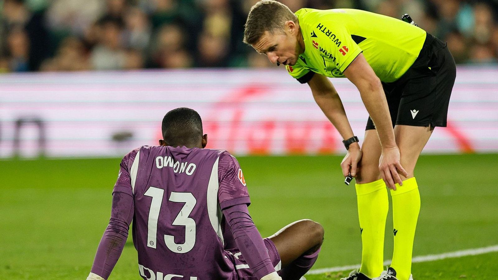 Hernández Hernández, con uniforme amarillo fluorescente, conversa con un jugador que lleva una camiseta morada con el número 13 y el nombre "OMONO", en un partido de fútbol.