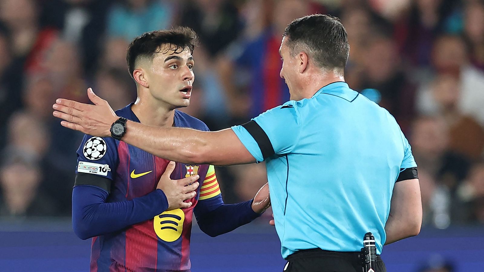 En un partido de la Champions League, un jugador con la camiseta azulgrana protesta una decisión arbitral. El árbitro, de espaldas, parece estar respondiendo a la protesta del jugador, que extiende su brazo.
