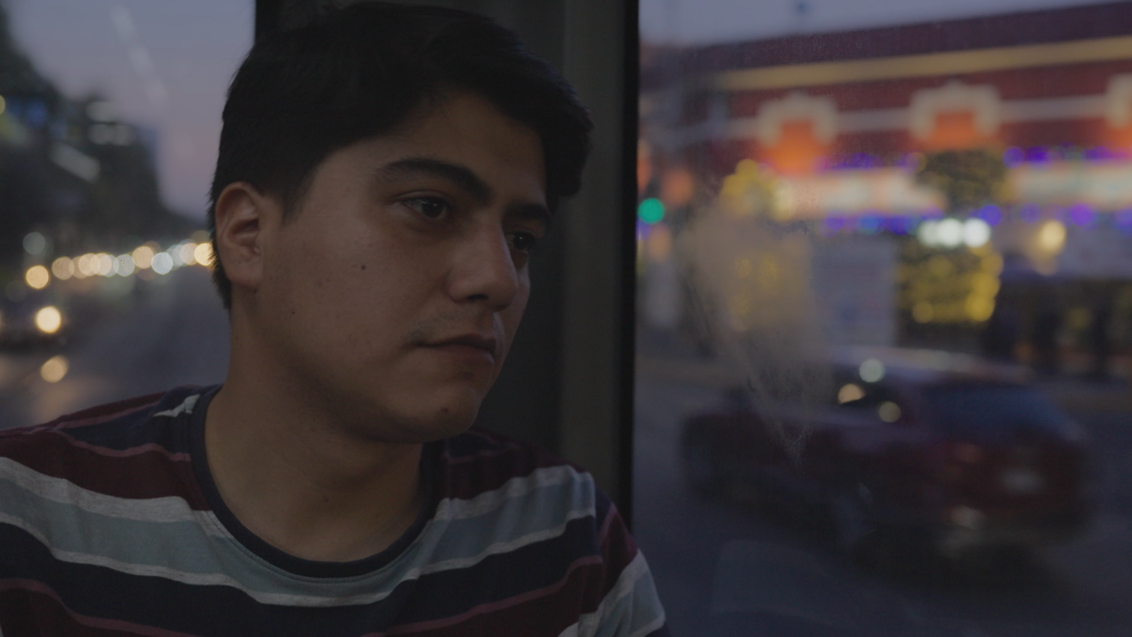 Un hombre joven, con camiseta a rayas, mira fijamente a través de una ventana. El reflejo en el cristal revela un paisaje urbano iluminado por la noche, con luces y un coche.