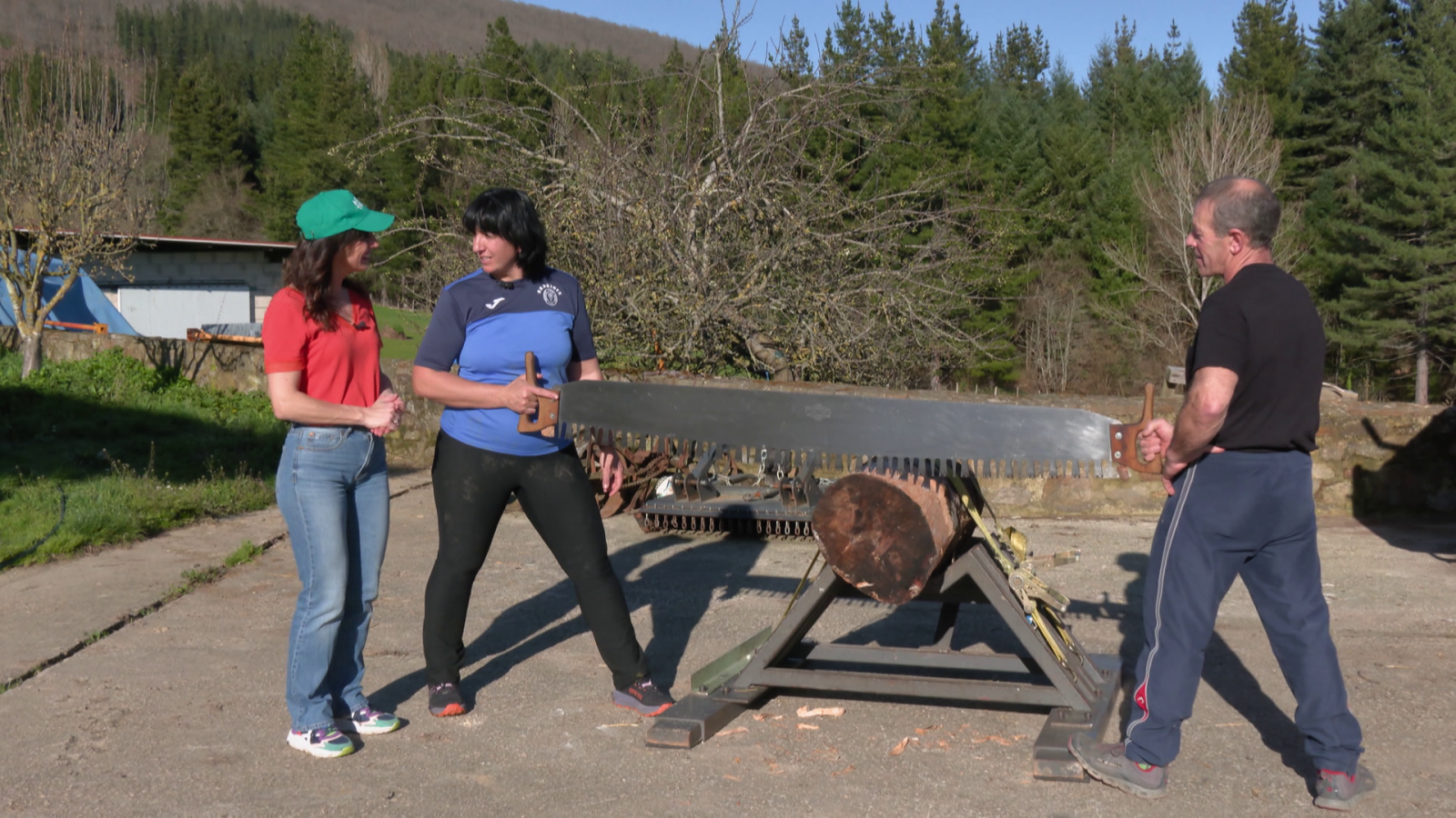 Garbiñe López de Uralde y un hombre trabajan juntos cortando un tronco con una sierra, mientras una mujer observa la escena. El tronco está sobre un soporte de madera, en un entorno con árboles y una casa.