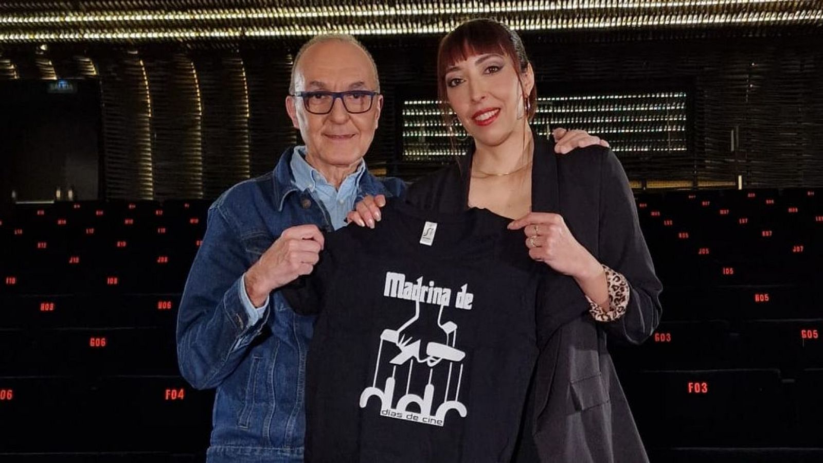 Dos personas posan en un espacio vacío, posiblemente un teatro. Ambos sostienen una camiseta negra con la inscripción "Madrina de" y una ilustración de una mano con hilos de marionetas, junto a la frase "días de cine".