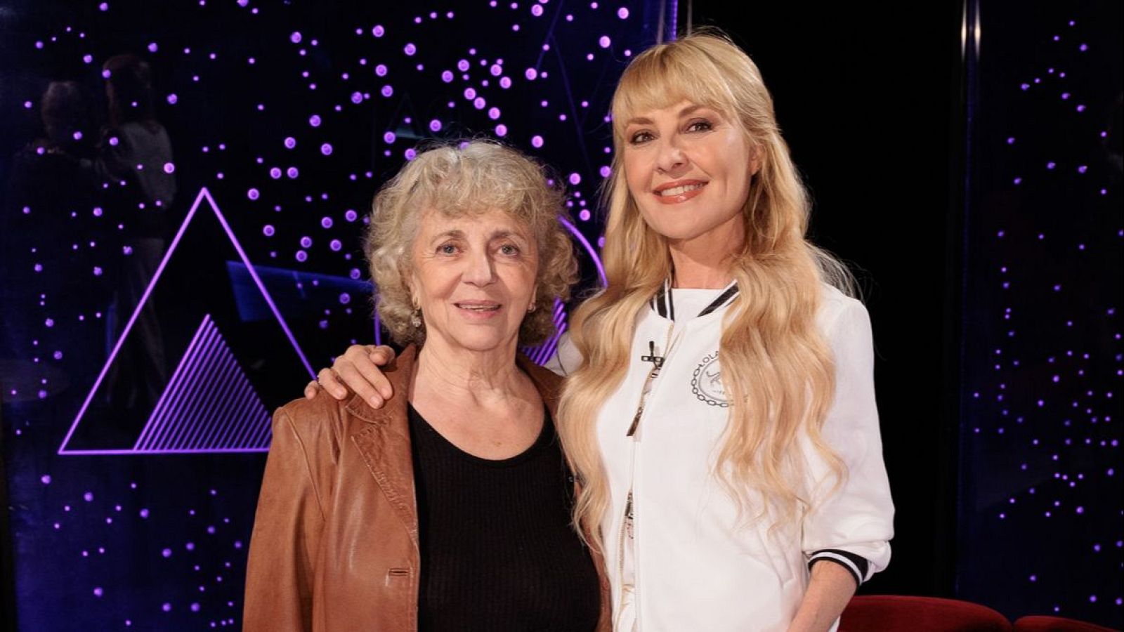 Dos mujeres posan juntas; una con cabello corto y rizado, vestida con ropa oscura y chaqueta de cuero, y otra con cabello largo y rubio, luciendo una chaqueta blanca con detalles en negro.