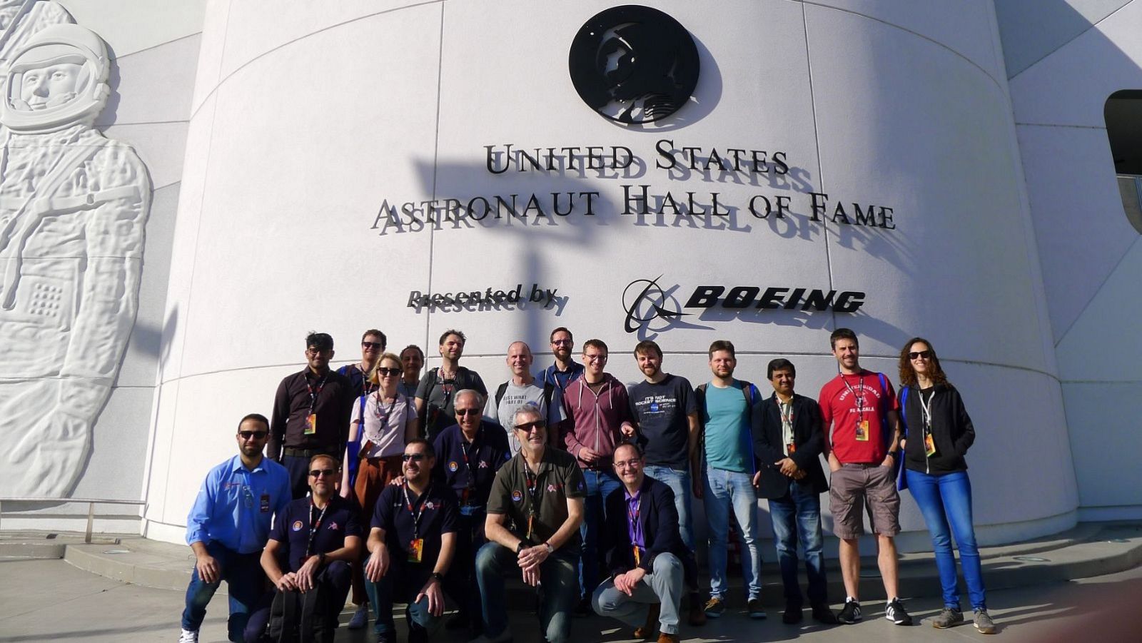 Un grupo diverso de personas se encuentra frente al edificio blanco con el nombre "UNITED STATES ASTRONAUT HALL OF FAME" y un relieve de astronauta, posiblemente miembros de un equipo de investigación.