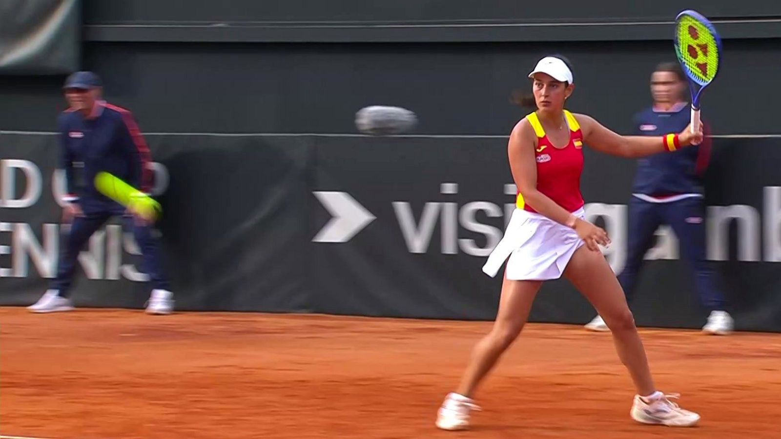 En una cancha de tierra batida, una tenista con camiseta roja y falda blanca se dispone a golpear la pelota con la raqueta en alto, frente a un juez de silla y público, con un panel publicitario al fondo.