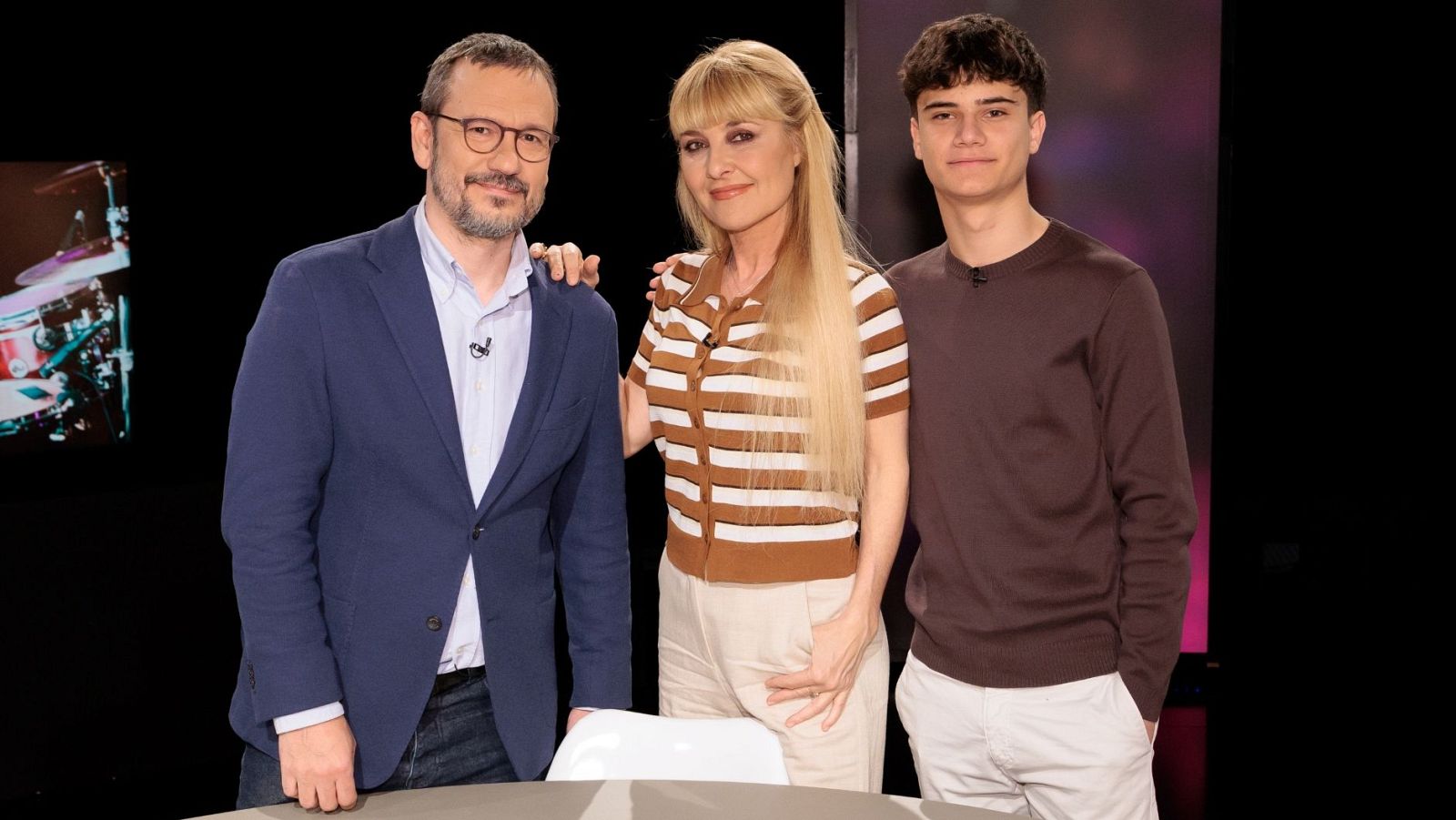 Tres personas posan frente a una mesa con mantel blanco; un hombre con gafas y chaqueta azul, una mujer con camisa de rayas y un joven con jersey marrón, con una pantalla borrosa de fondo.