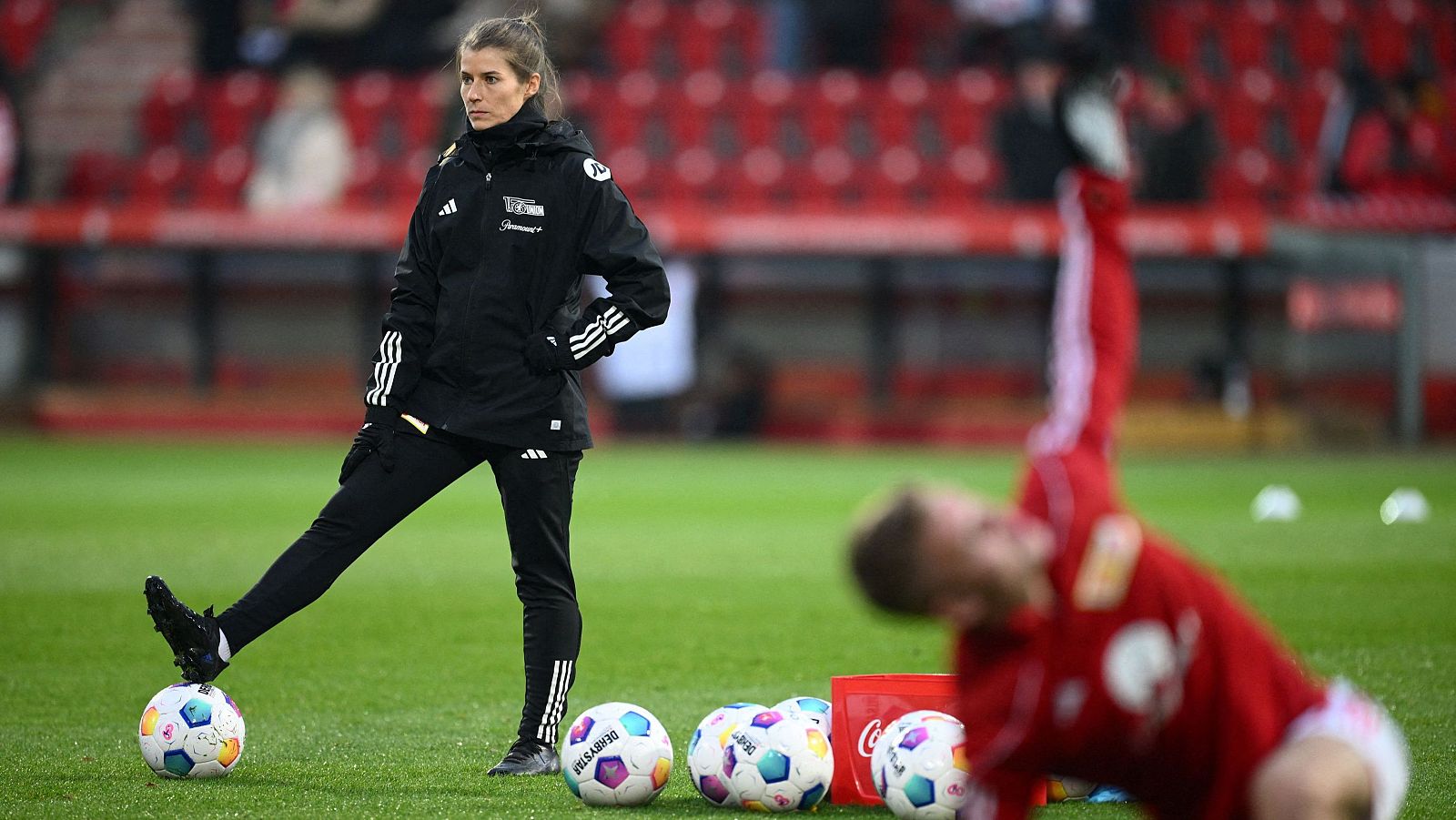 Marie-Louise Eta, entrenadora asistente, posa en un campo de fútbol con ropa deportiva negra, incluyendo una chaqueta con el logo del equipo y las tres rayas de Adidas. Hay balones y una bolsa roja en primer plano, y una persona estirándose al fondo.