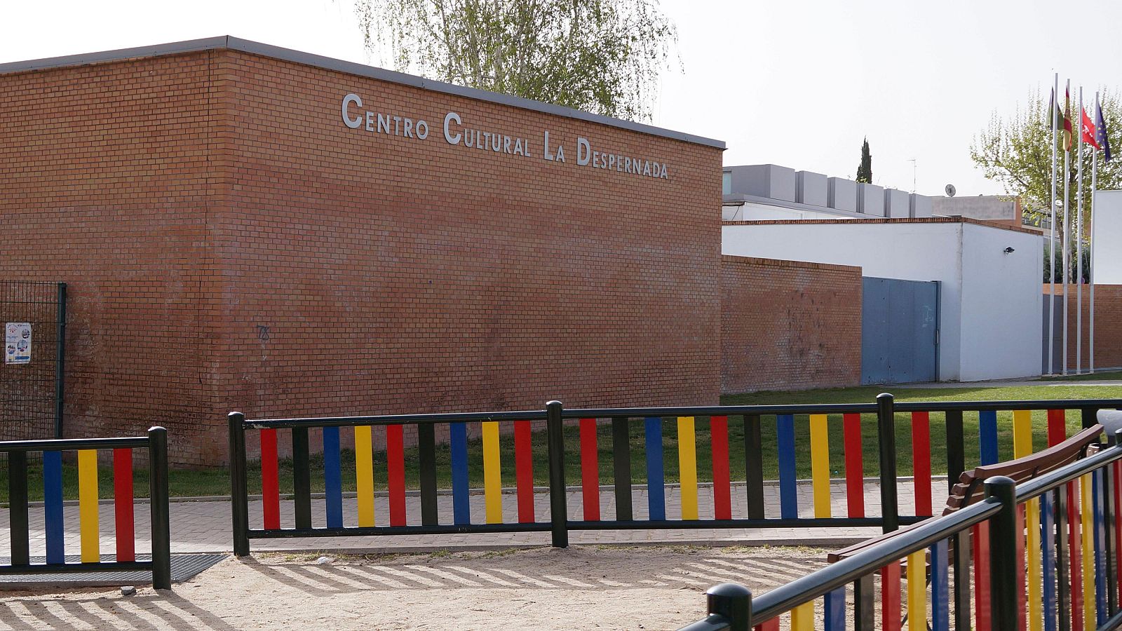 Un edificio de ladrillo con el letrero "CENTRO CULTURAL LA DESPERNADA" se encuentra frente a una valla de colores. Al fondo, se aprecian otros edificios y una puerta azul, con banderas ondeando a un lado.