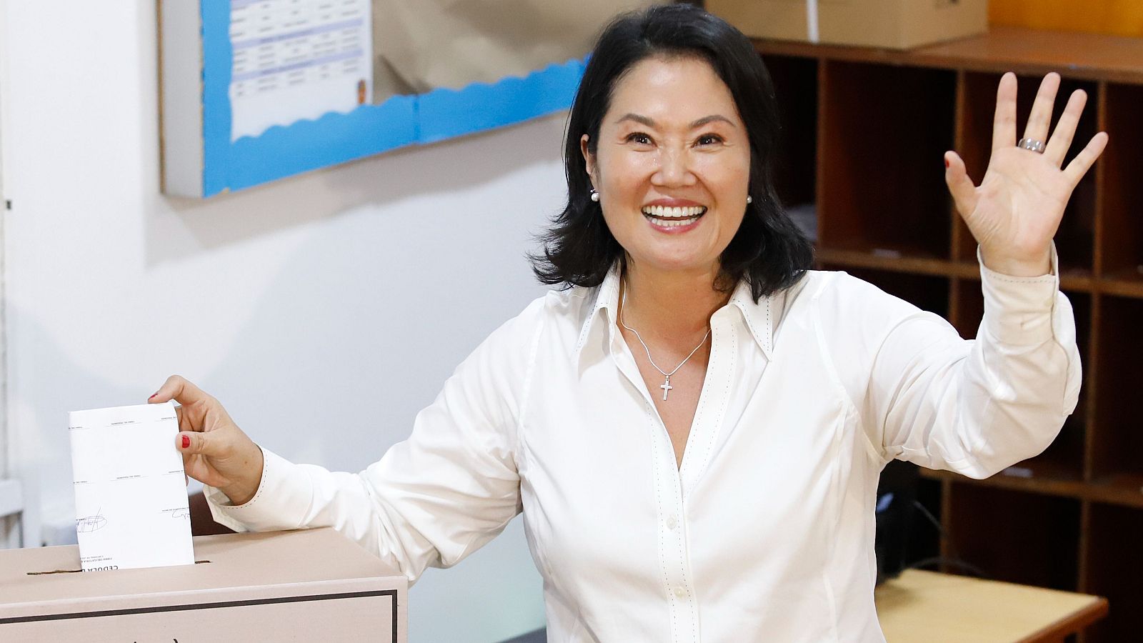 En una urna electoral, Keiko Fujimori, candidata a la presidencia, deposita su voto. Viste una camisa blanca y un collar con una cruz, mientras sonríe y saluda.