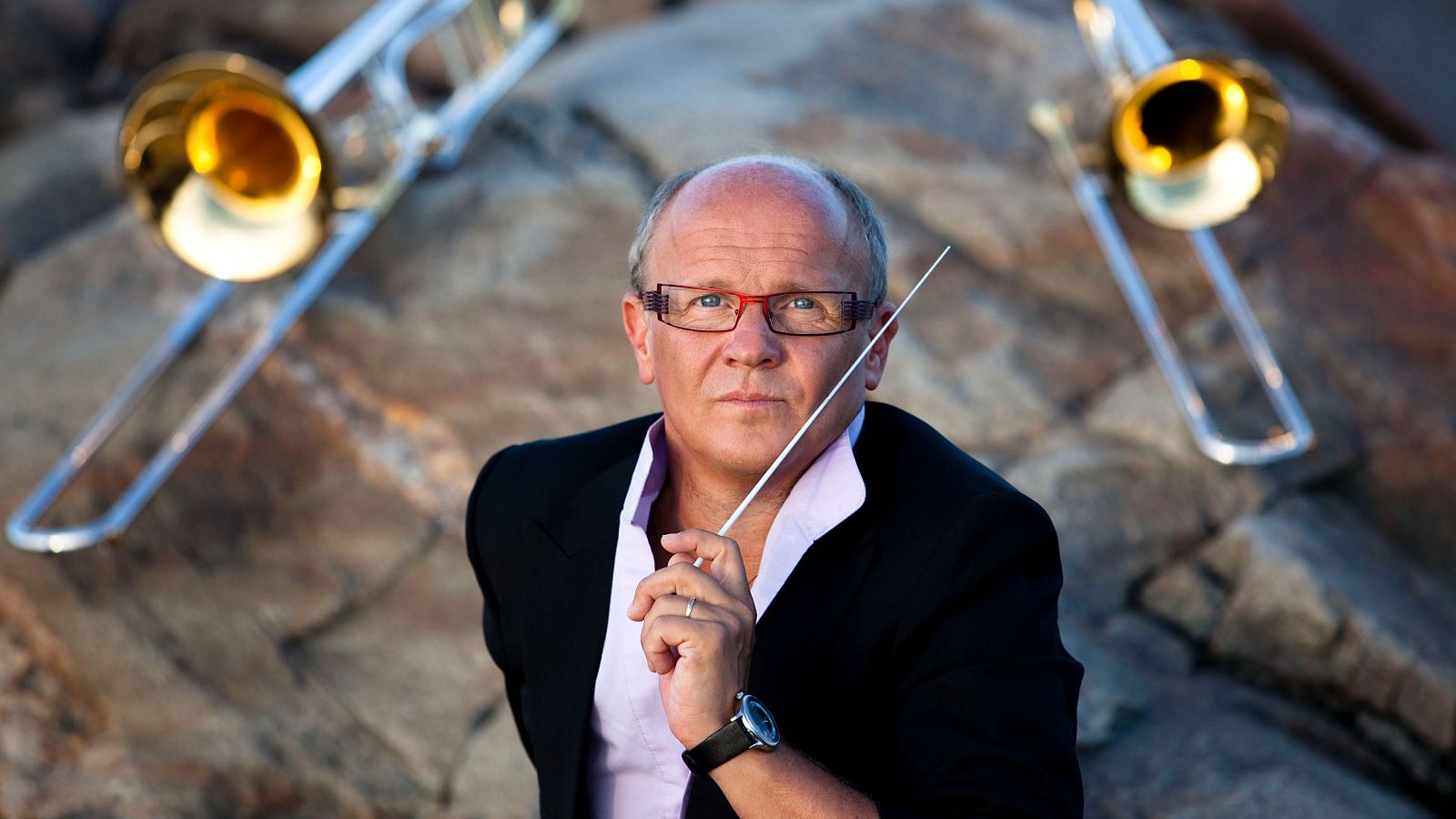 Christian Lindberg, con gafas y camisa clara, dirige con una batuta. Detrás de él, sobre un fondo rocoso, se aprecian dos trombones.