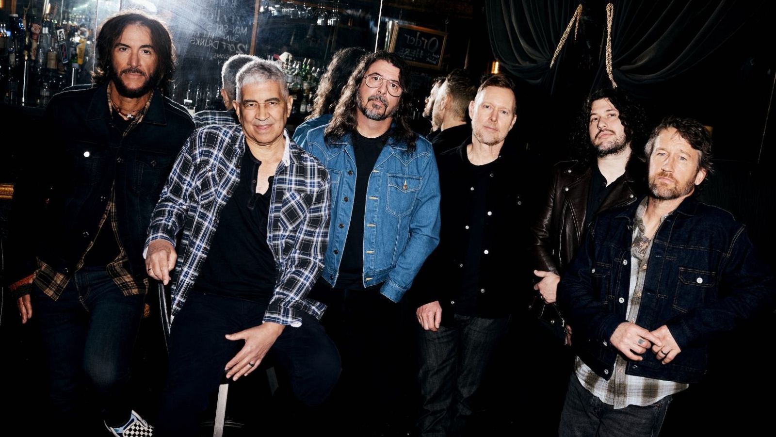 Un grupo de músicos posa en un bar con iluminación tenue. Los hombres, con diferentes estilos de cabello y barba, visten chaquetas y camisas a cuadros, creando una atmósfera informal y relajada.