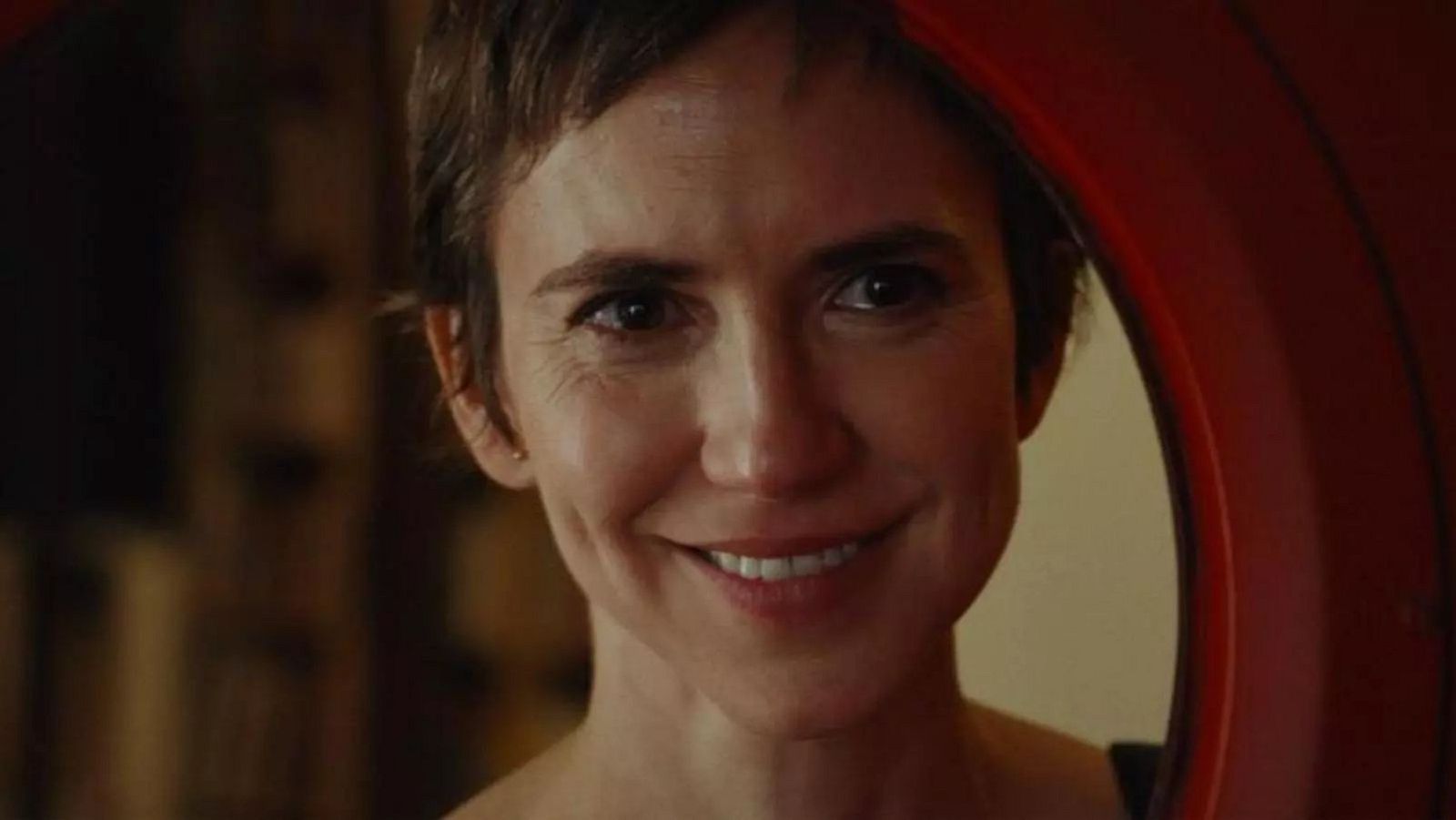 En primer plano, Aina Clotet sonríe con el pelo corto castaño, enmarcada parcialmente por un elemento circular rojo, con un fondo borroso que sugiere un interior.