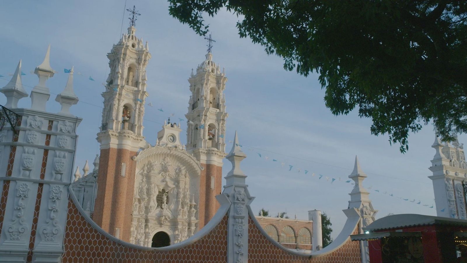 Una iglesia con torres de ladrillo rojo y fachada blanca, decorada con detalles arquitectónicos y esculturas, se alza frente a una valla ornamental. El cielo parcialmente nublado y la vegetación enmarcan la escena.